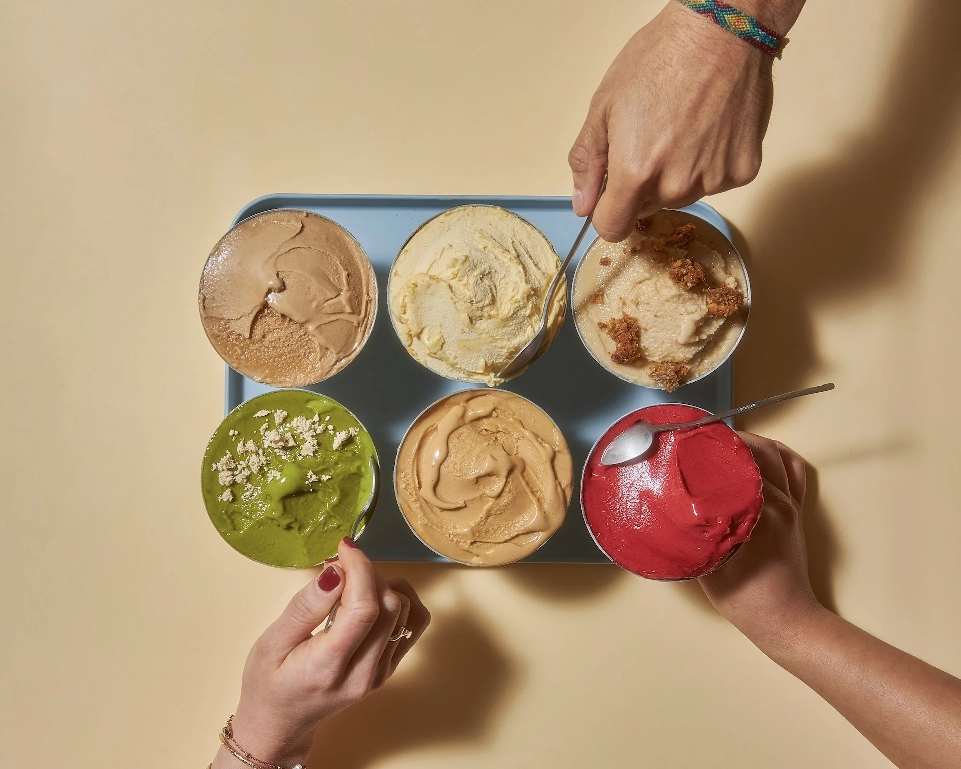 Six pots de glace de différentes saveurs, avec trois mains tenant des cuillères, sur une table beige.