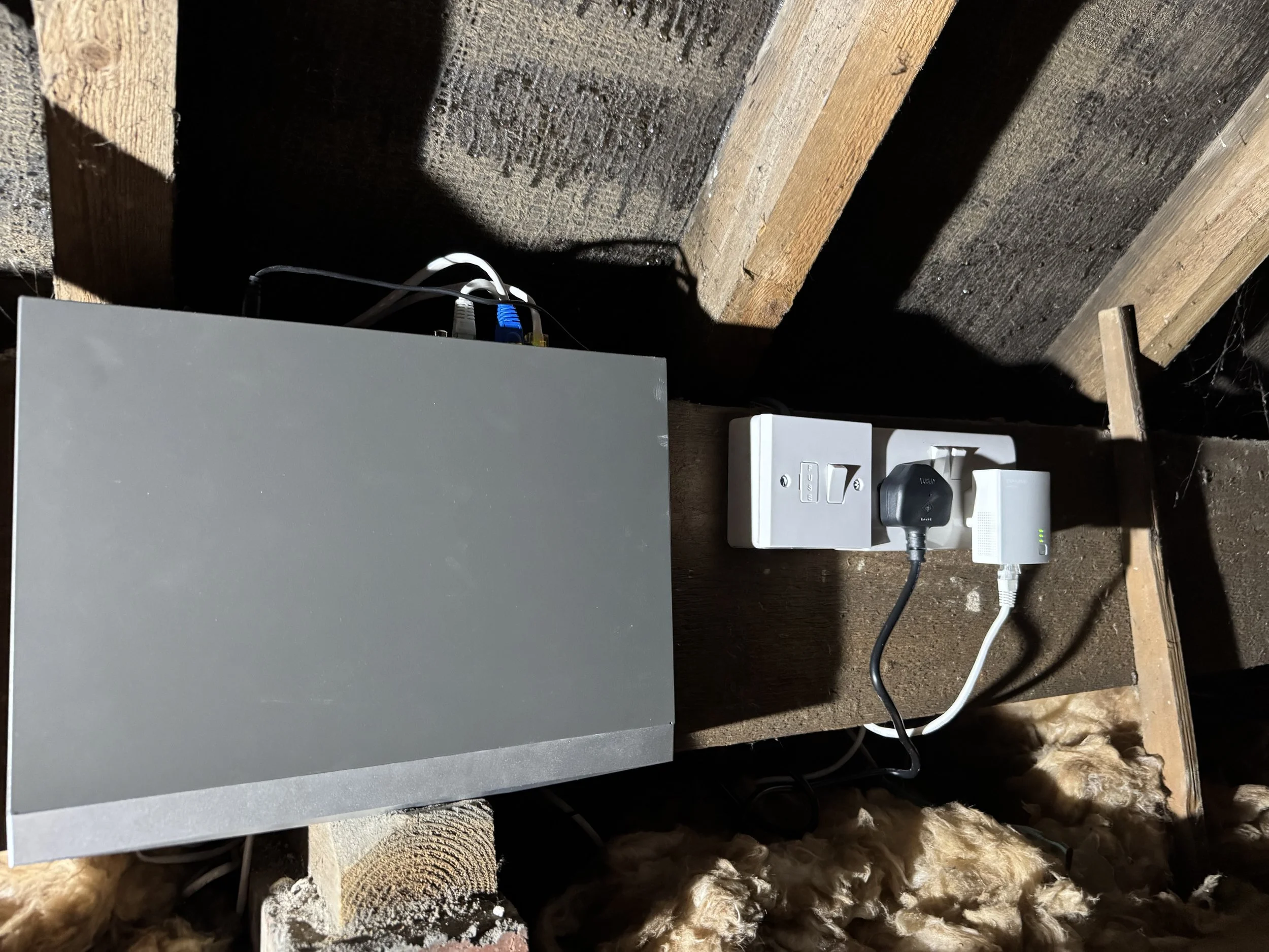 A gray electrical box connected to white and black power cords, plugged into a wall outlet, with an unfinished wooden wall and insulation at the bottom of the image.