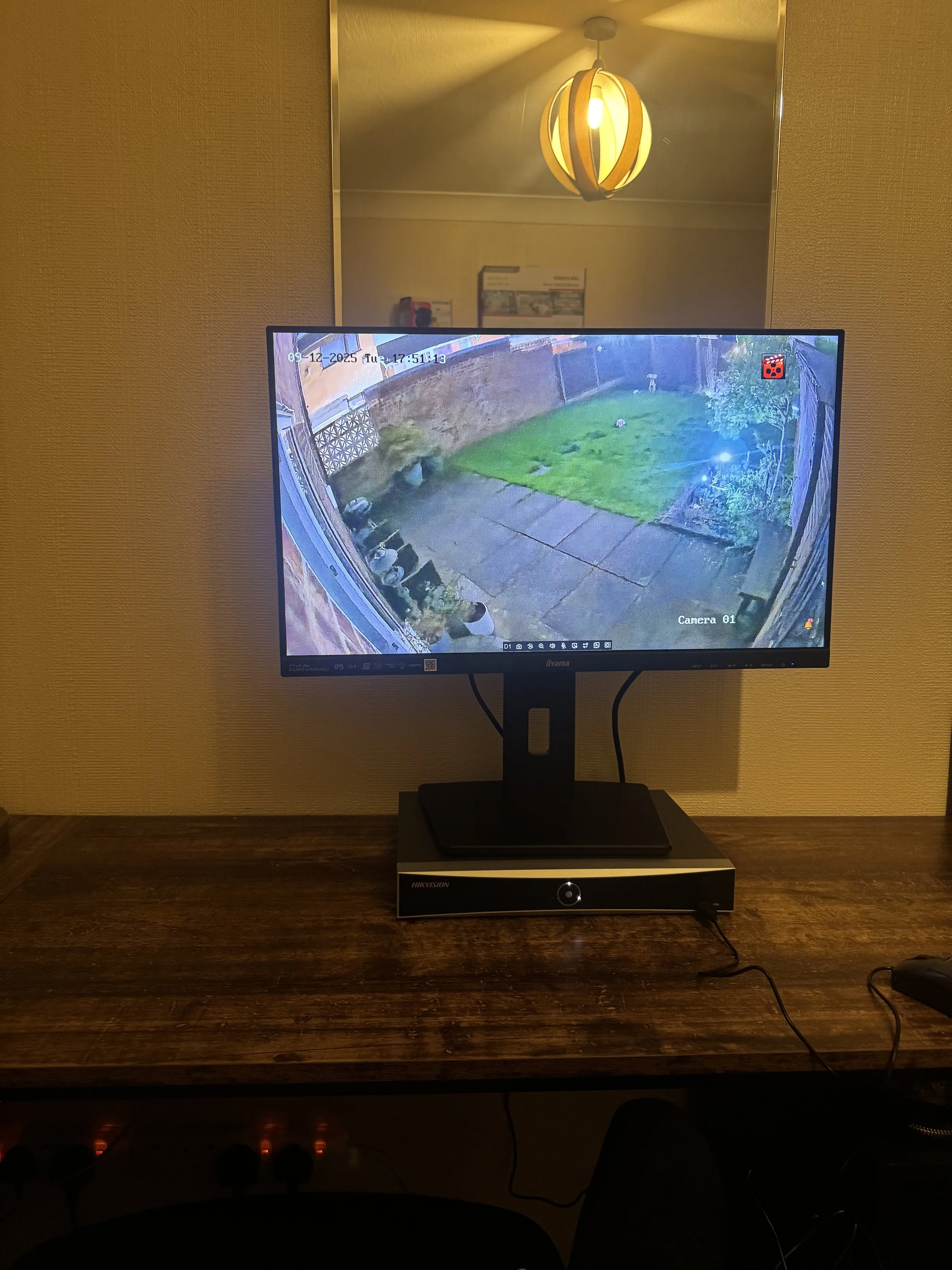 Security monitor displaying a backyard scene with grass, concrete path, and plants, mounted above a black device on a wooden desk.