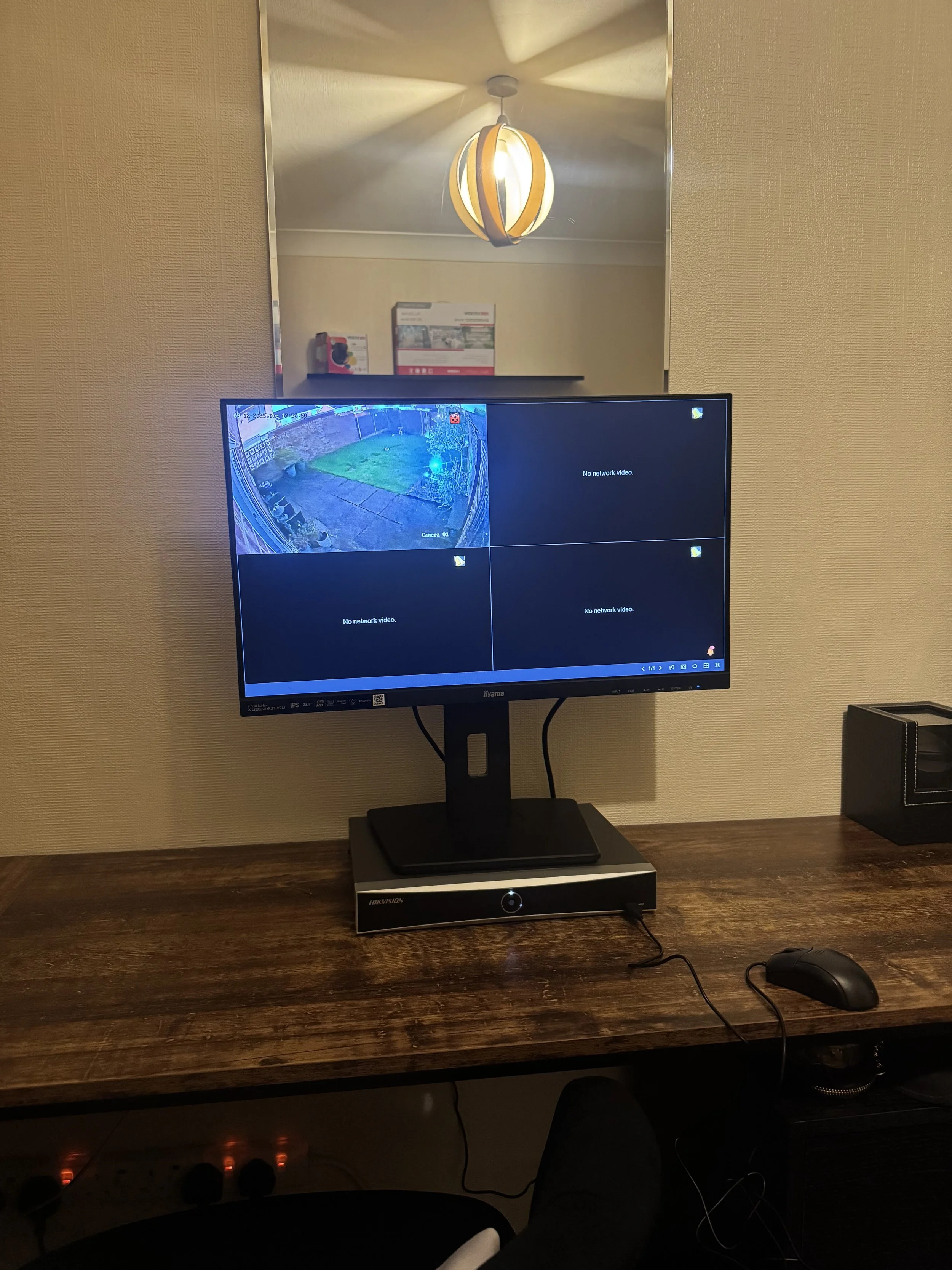 Computer monitor displaying surveillance camera feed and network status screens. The monitor is on a wooden desk with a mouse, in a room with a mirror, mirror reflection shows a ceiling light and boxes in the background.