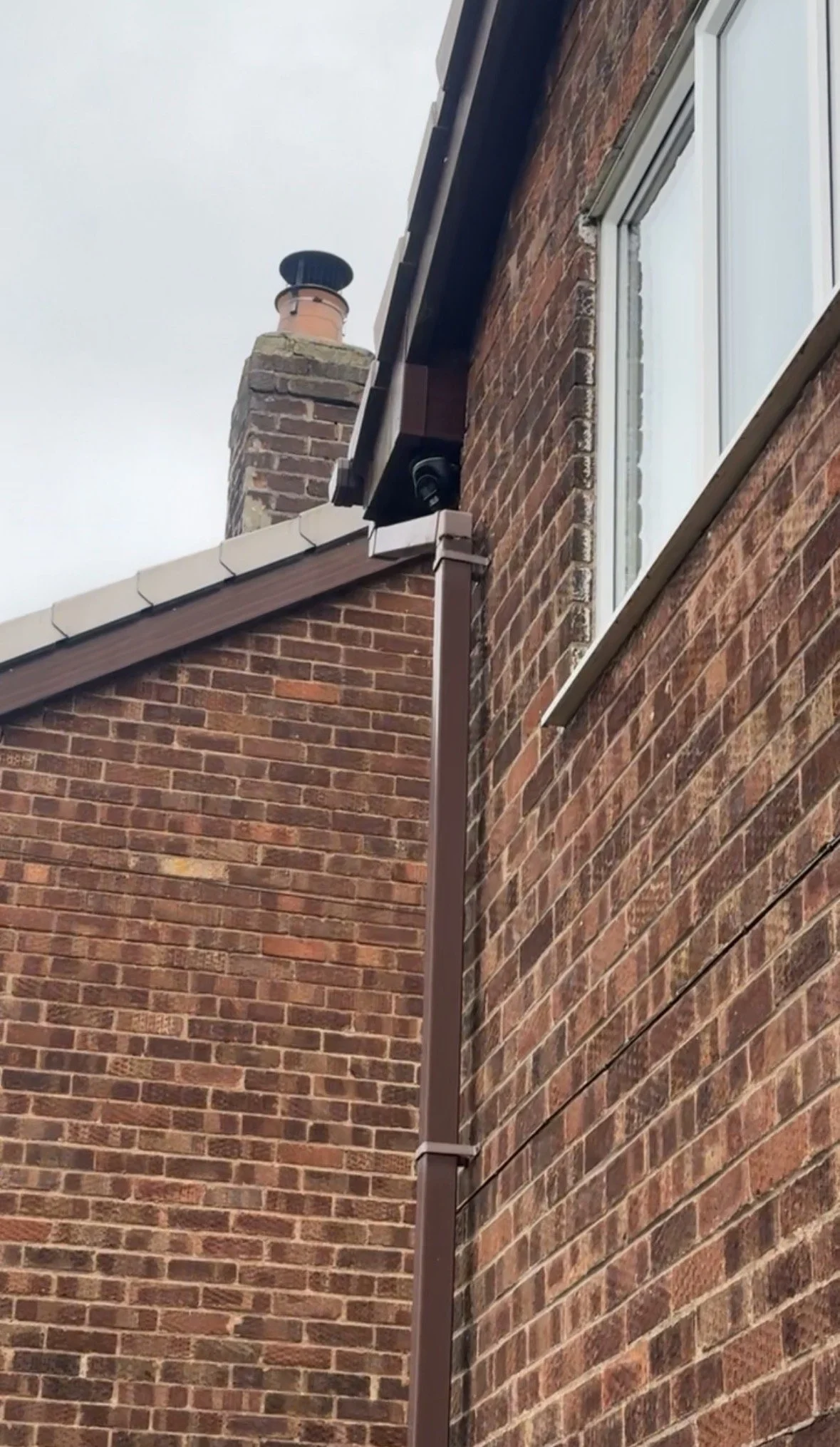Close-up of the corner of a brick house showing a black security camera near a window, with a brown downspout running along the side.