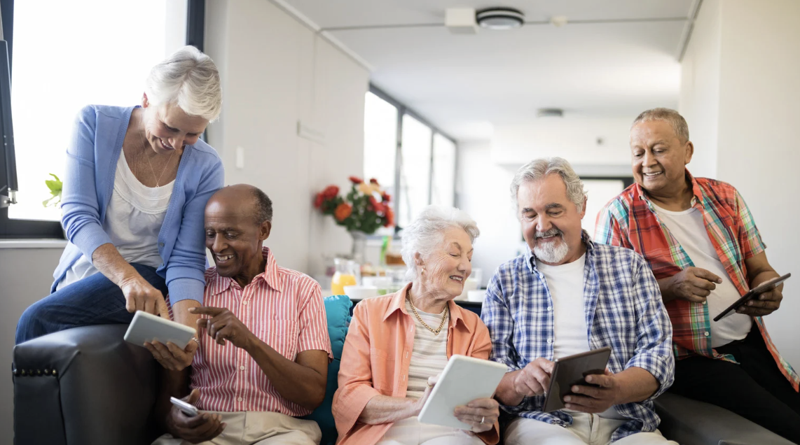 Six seniors gathered in a bright living room, some holding tablets, smiling, and interacting with each other.