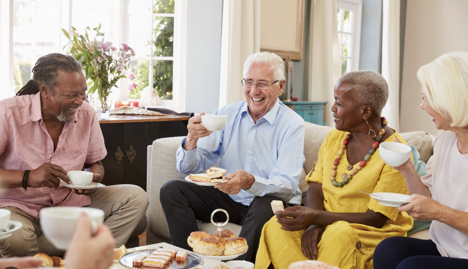 Group of five elderly friends sitting on a couch, enjoying tea and snacks, smiling and engaging in conversation in a bright living room.