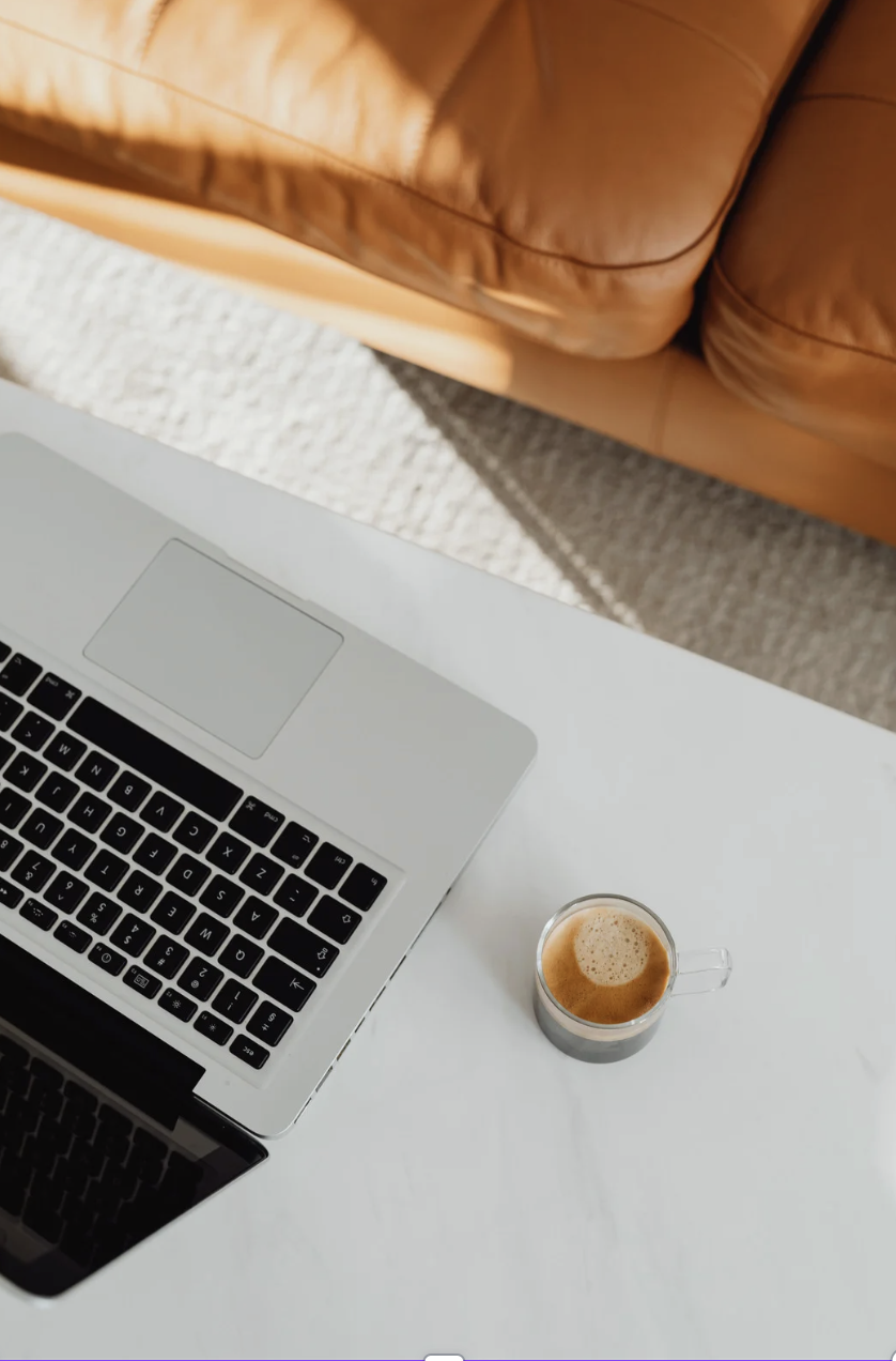 A silver laptop with a black keyboard on a white desk, next to a glass mug of coffee, with a brown leather couch in the background.