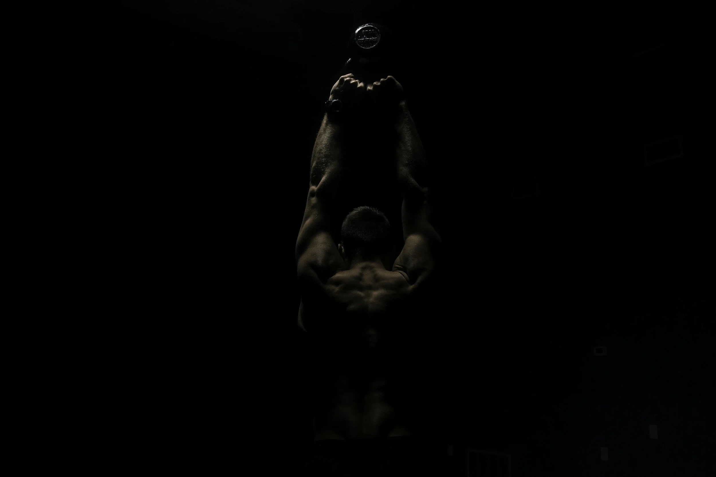 A person performing a headstand pose, facing downward with their arms supporting their body and legs extended upright, in a dark environment.