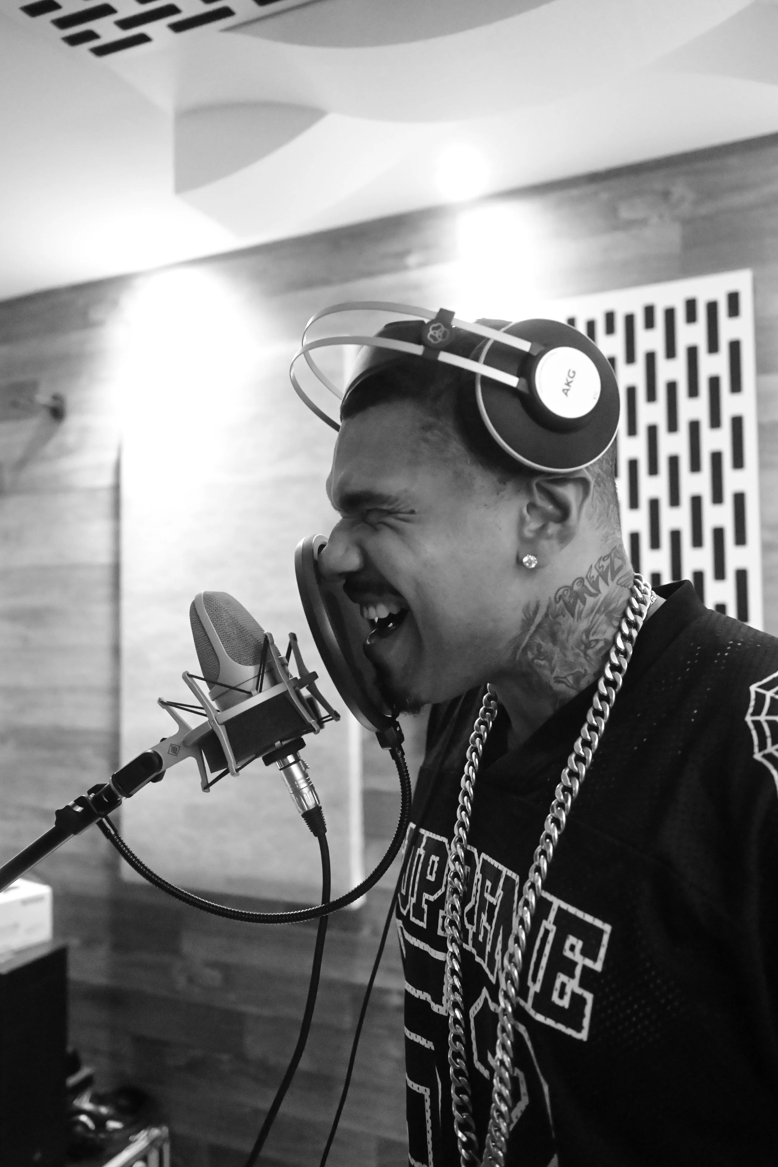 Homem com tatuagens, usando cadeado no pescoço, ouvindo música com fones de ouvido AKG, cantando em estúdio de gravação.