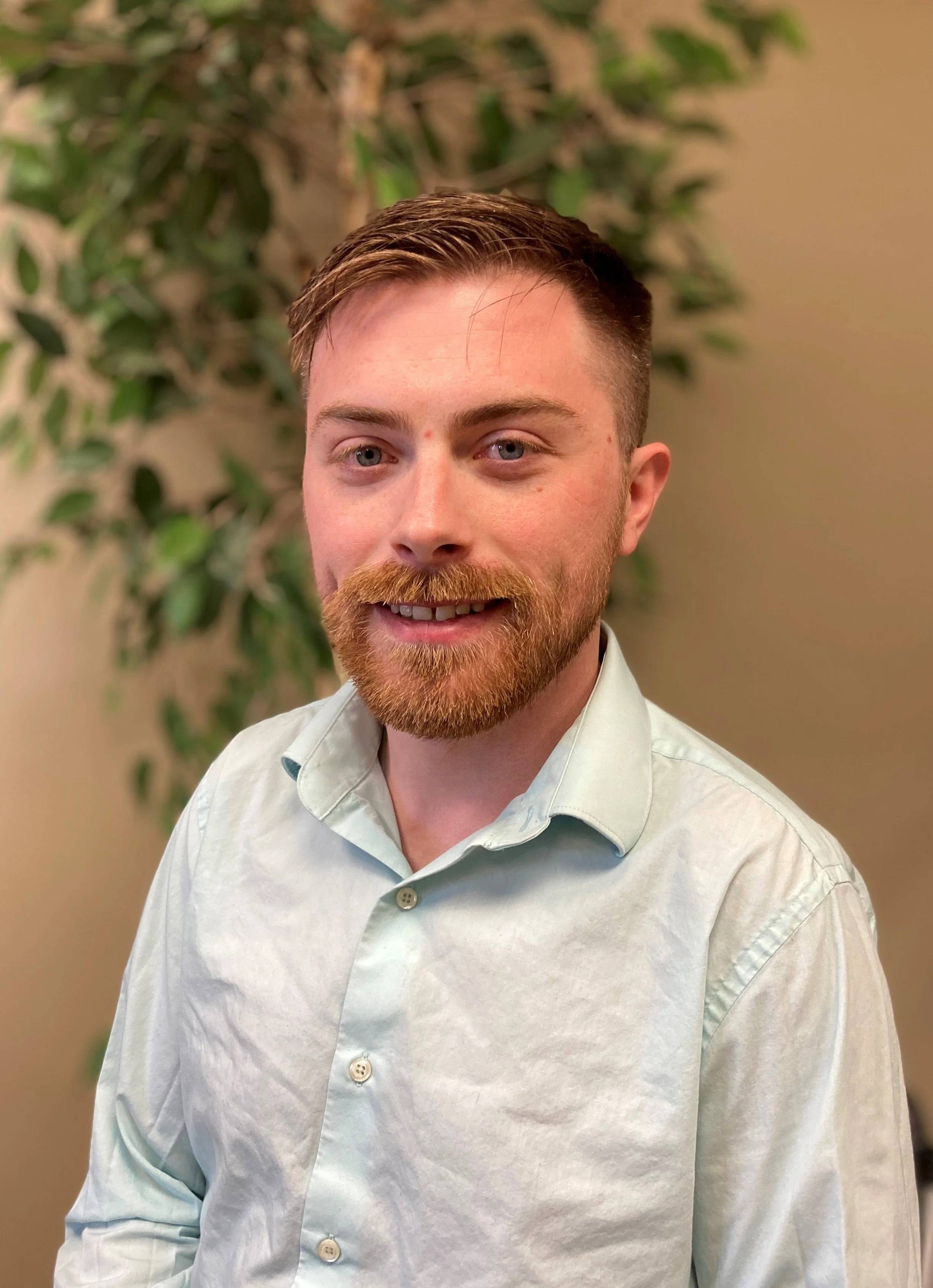 A young man with light skin, reddish-brown hair, a beard, and blue eyes, smiling while wearing a white dress shirt. There is a green leafy plant behind him and a beige wall in the background.