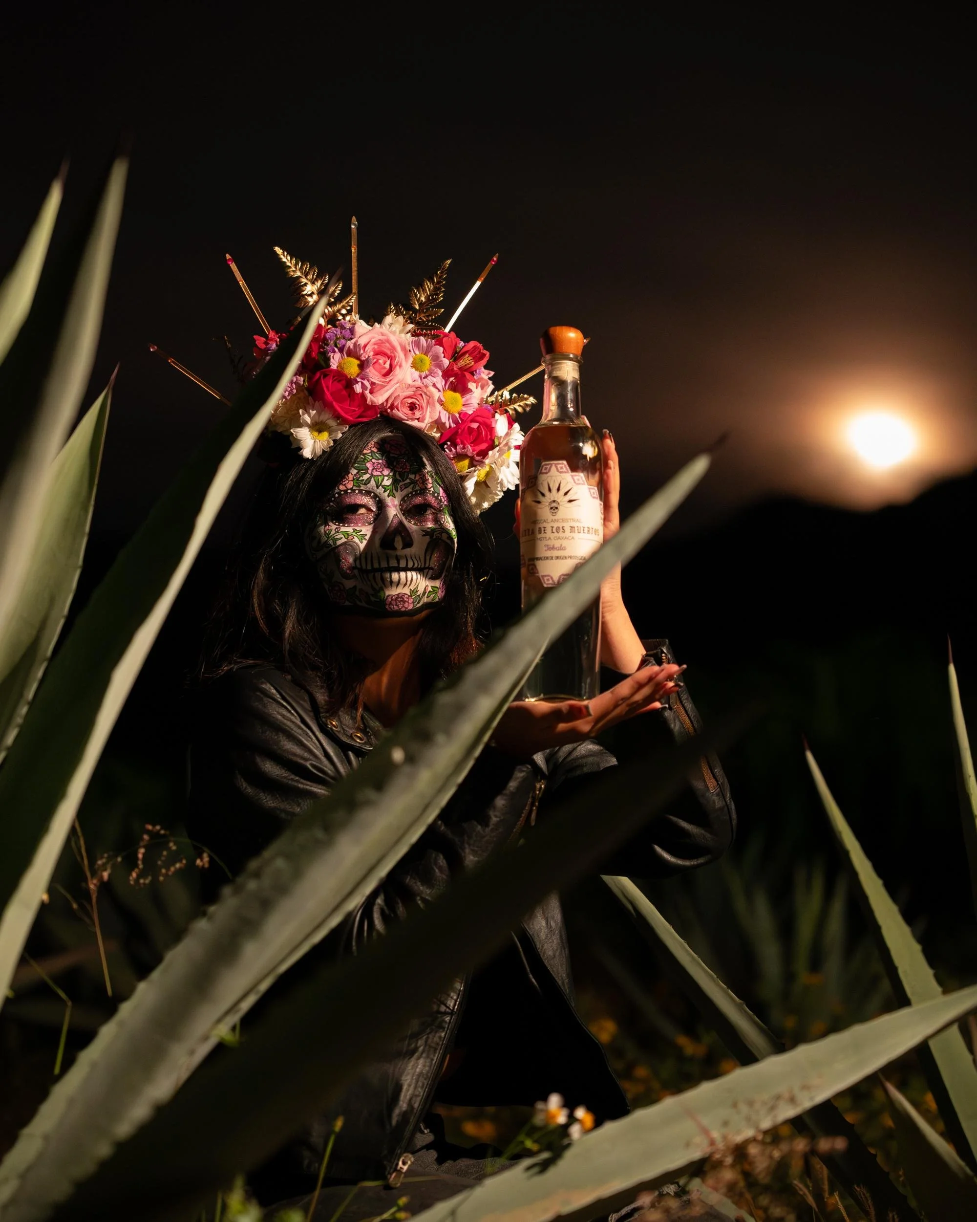 Person dressed in a leather jacket wearing a skull face paint and a colorful flower crown, holding a bottle, surrounded by agave plants during nighttime with the moon visible in the sky.