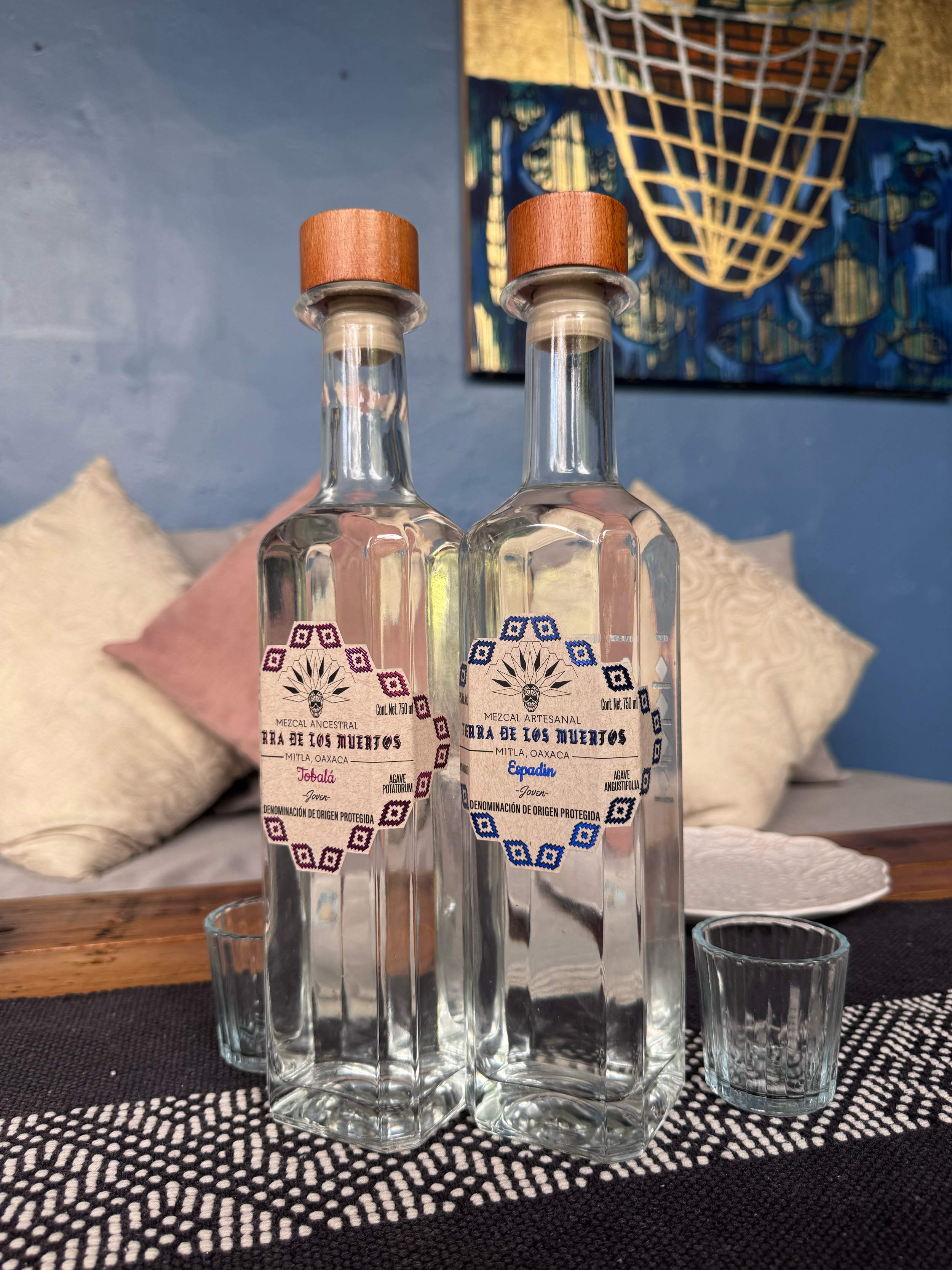Two tall glass bottles with wooden caps and illustrated labels, placed on a table with small glasses, in a room with blue wall and colorful artwork.