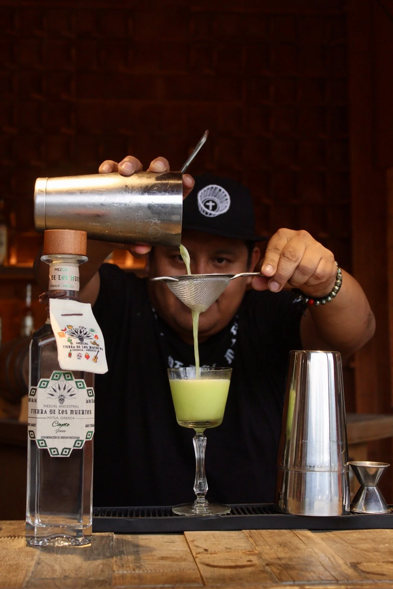 Bartender pouring a green cocktail through a strainer into a glass, with a bottle of mezcal nearby.