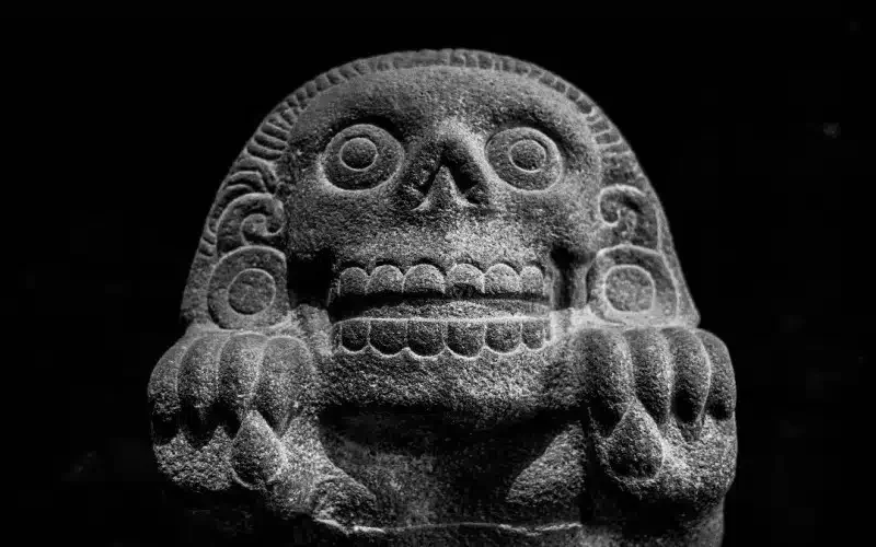 Stone sculpture of a stylized skull with large round eye sockets, a nasal opening, and teeth, set against a black background.