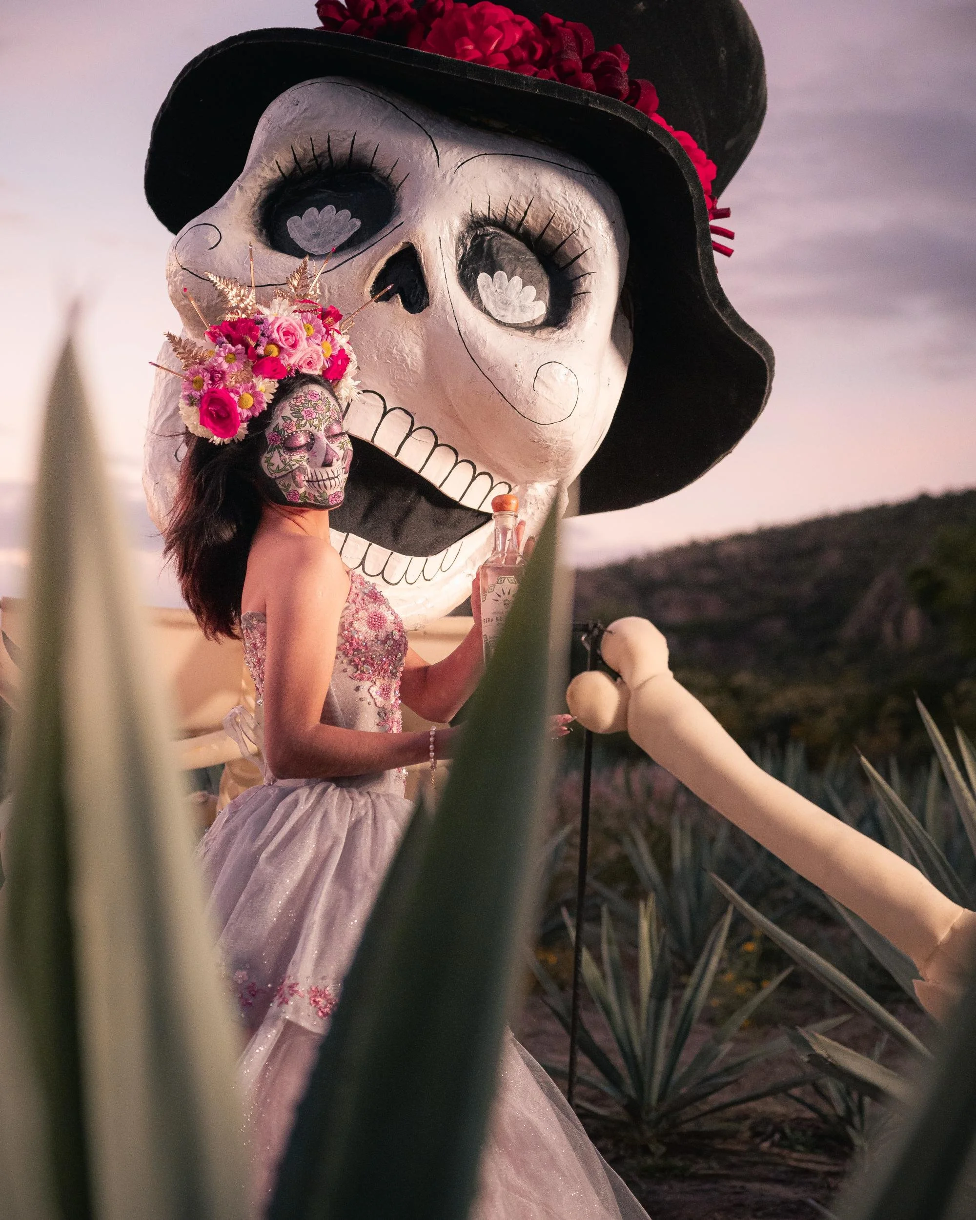 A woman dressed in a floral gown with face paint resembling a sugar skull, wearing a large flower crown, standing in front of a large decorative skull with a hat, in an outdoor setting during sunset.