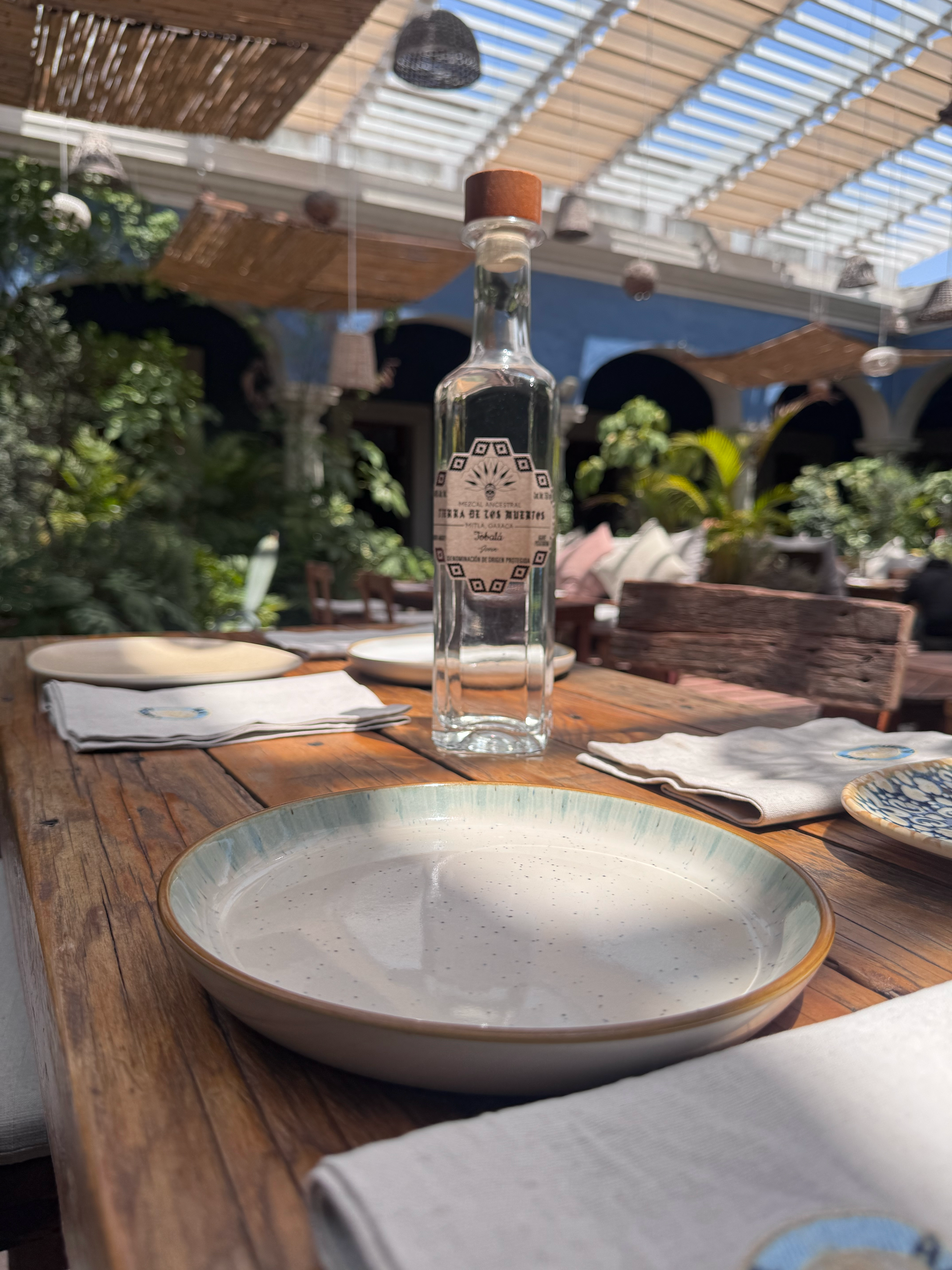 A rustic wooden dining table set with a ceramic plate, cloth napkins, and a glass bottle of mezcal, in an outdoor restaurant with a pergola overhead and lush greenery in the background.