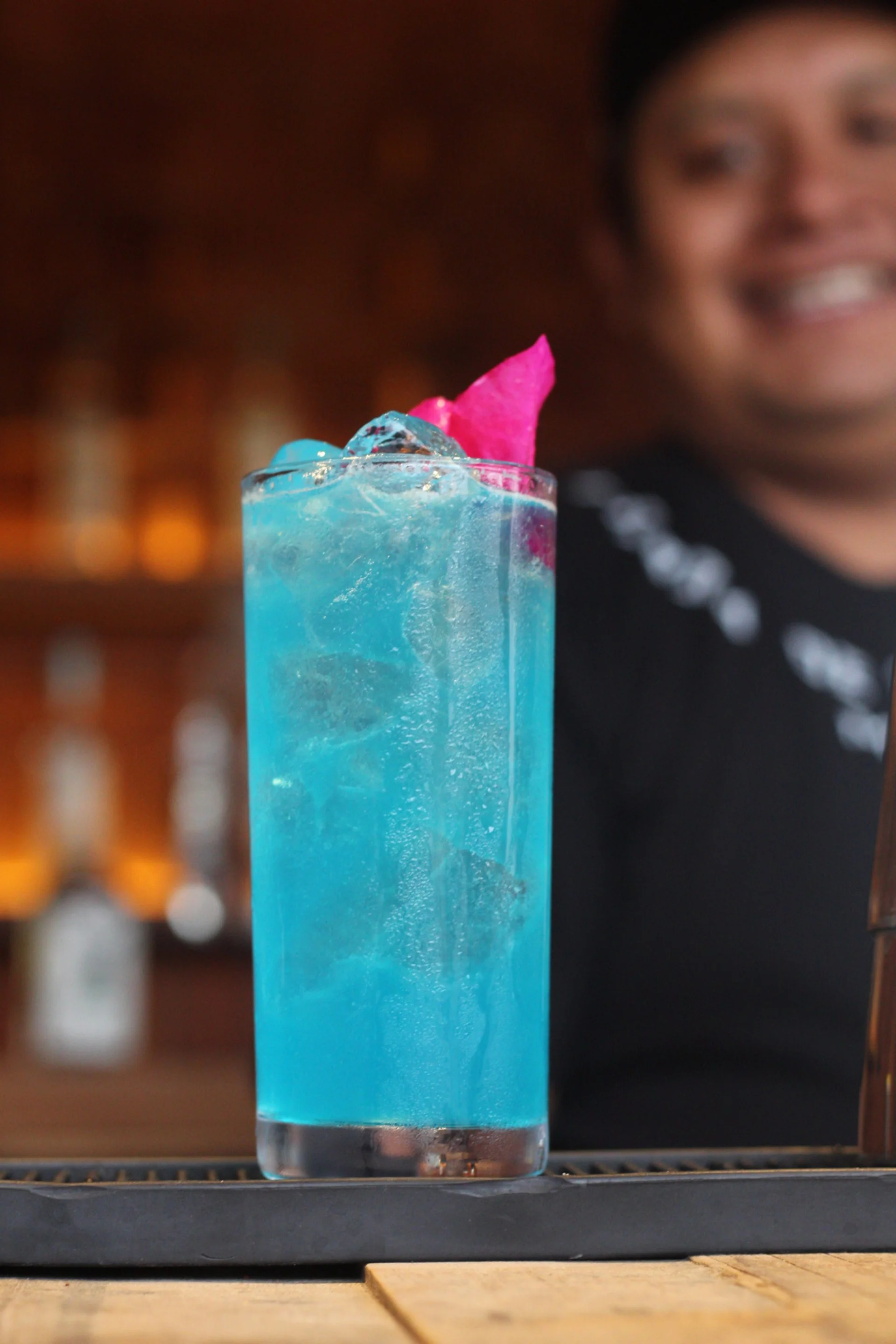 A tall glass of blue cocktail with ice and a pink garnish on top, placed on a black bar mat with a blurred smiling person in the background.