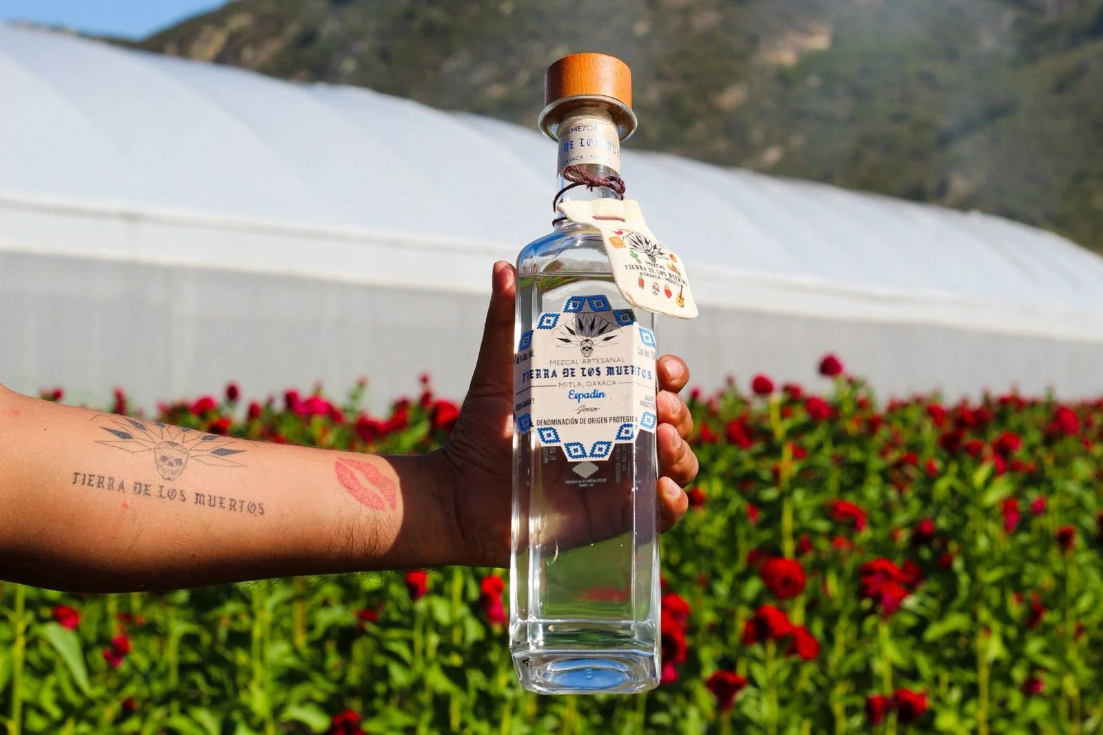 A hand with tattoos holding a clear glass bottle of mezcal with a wooden cap and decorative tags, standing outdoors in front of a garden with red flowers and a white greenhouse in the background.
