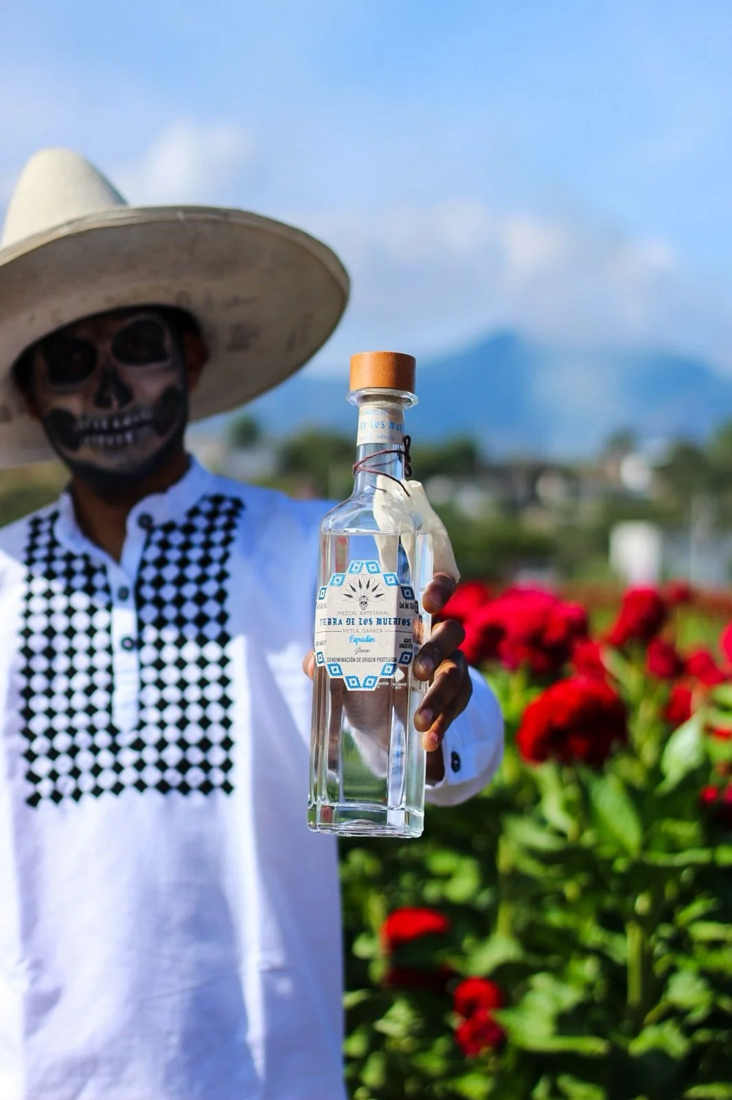 A person dressed in traditional Mexican clothing with a skull face design and wearing a large sombrero, holding a clear bottle of tequila in front of red flowers and greenery.