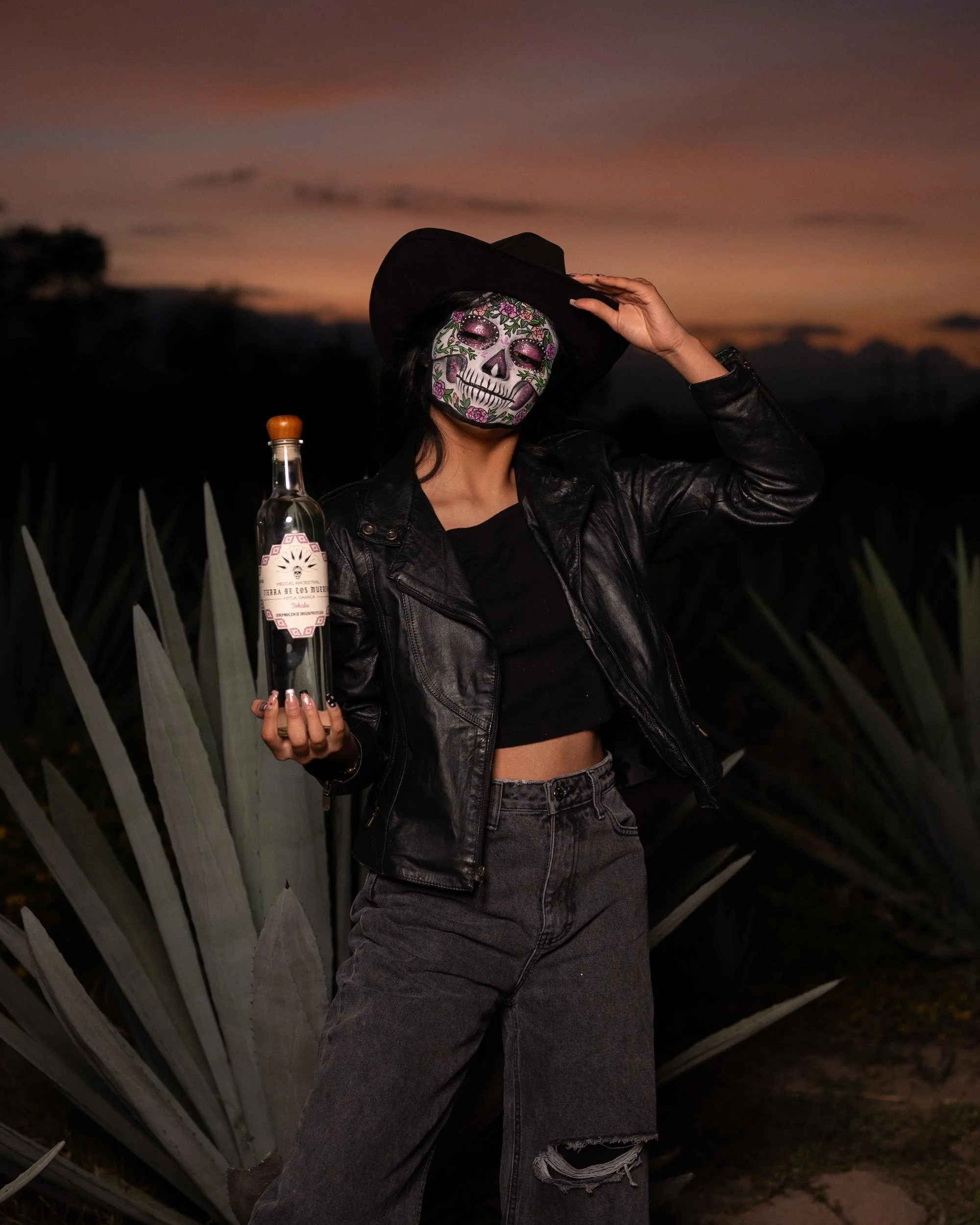 A woman wearing a skeleton face mask, black leather jacket, black crop top, and ripped jeans, holding a bottle, standing outdoors during sunset with agave plants in the background.