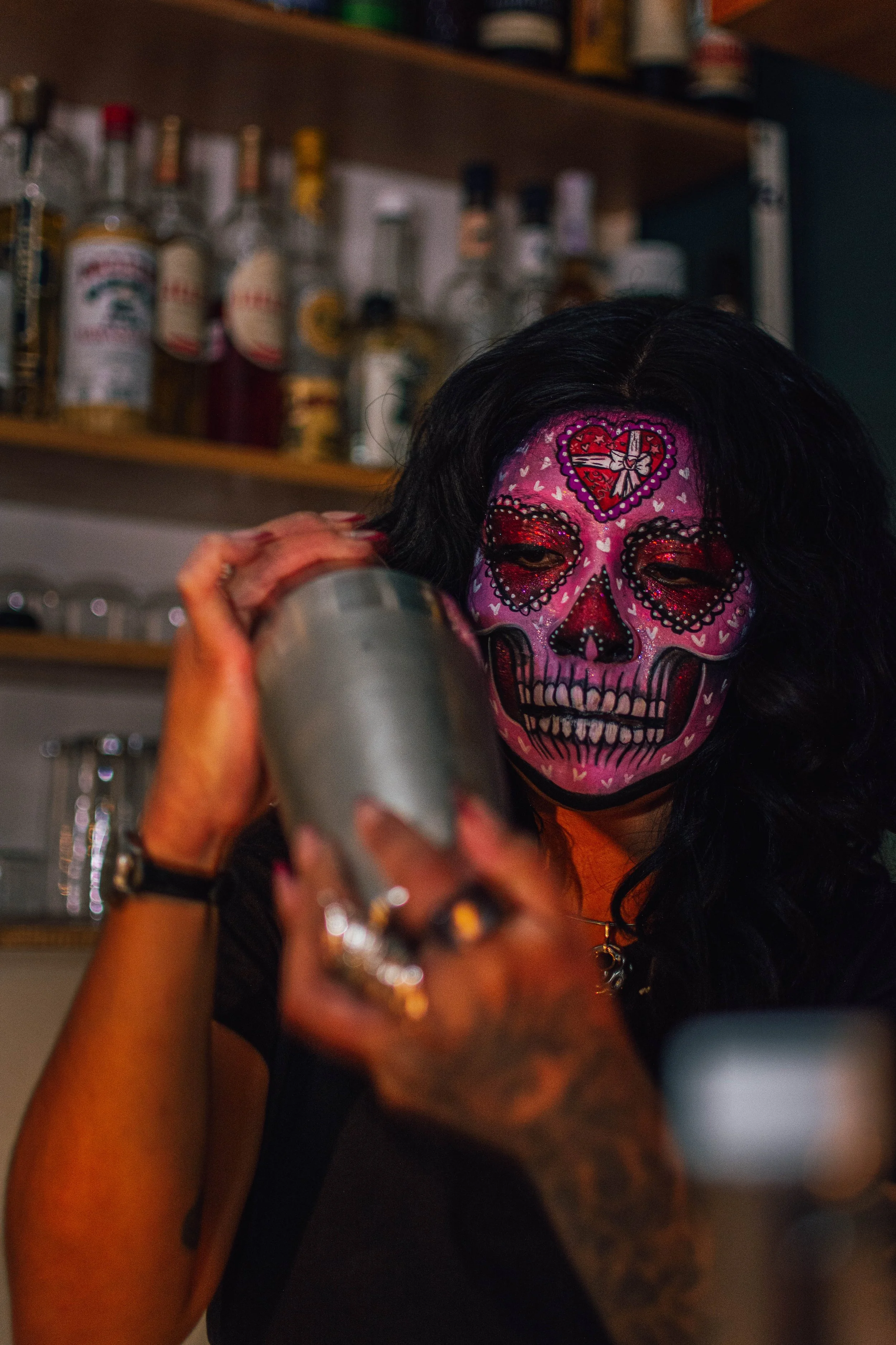 A woman with face paint resembling a sugar skull, with pink, black, and red designs, is shaking a cocktail shaker in a bar or restaurant setting, with shelves of bottles behind her.