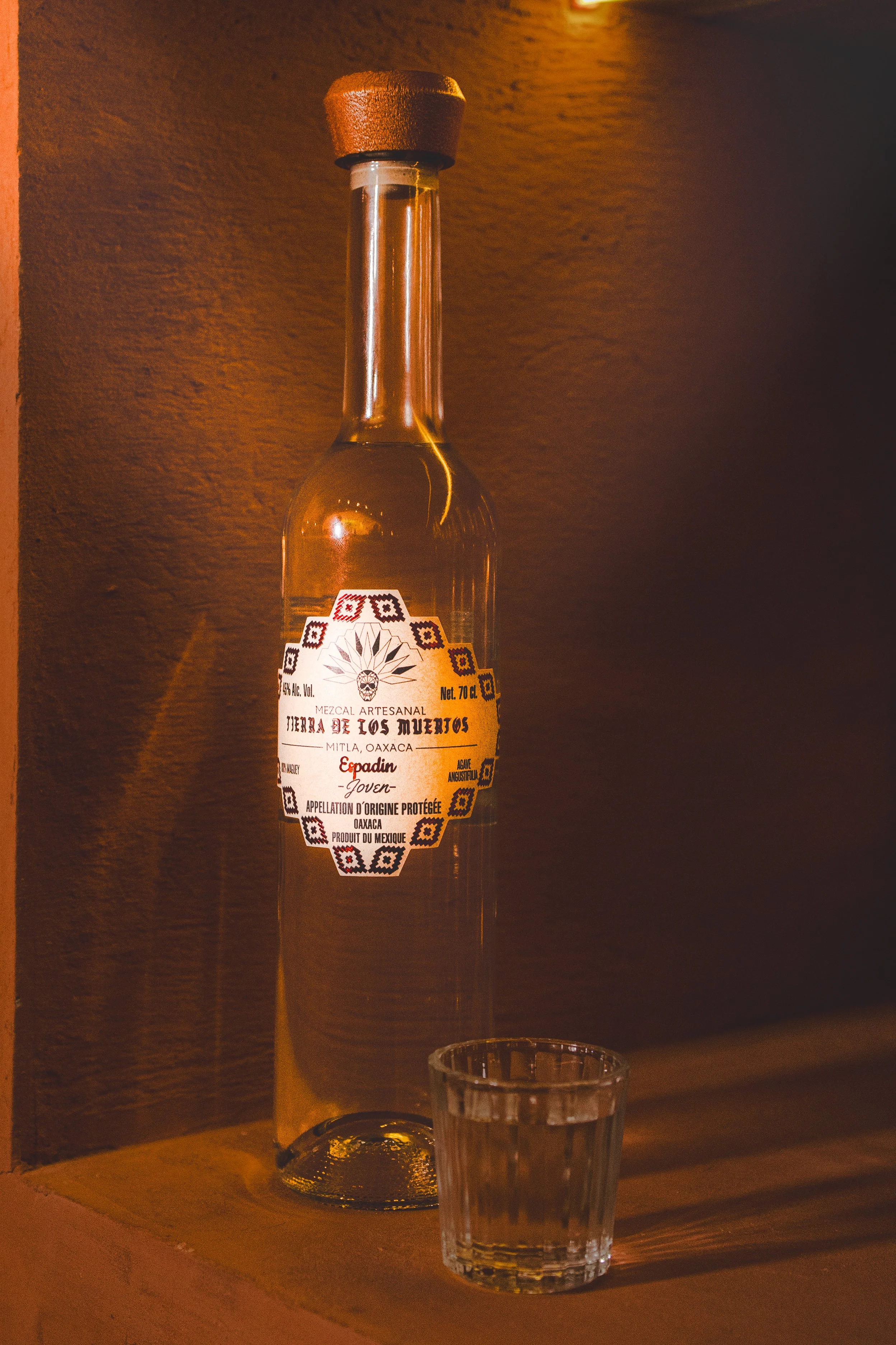 A tall glass bottle of mezcal with a paper label, and a small empty shot glass placed in front of it on a wooden surface against a warm-colored wall.