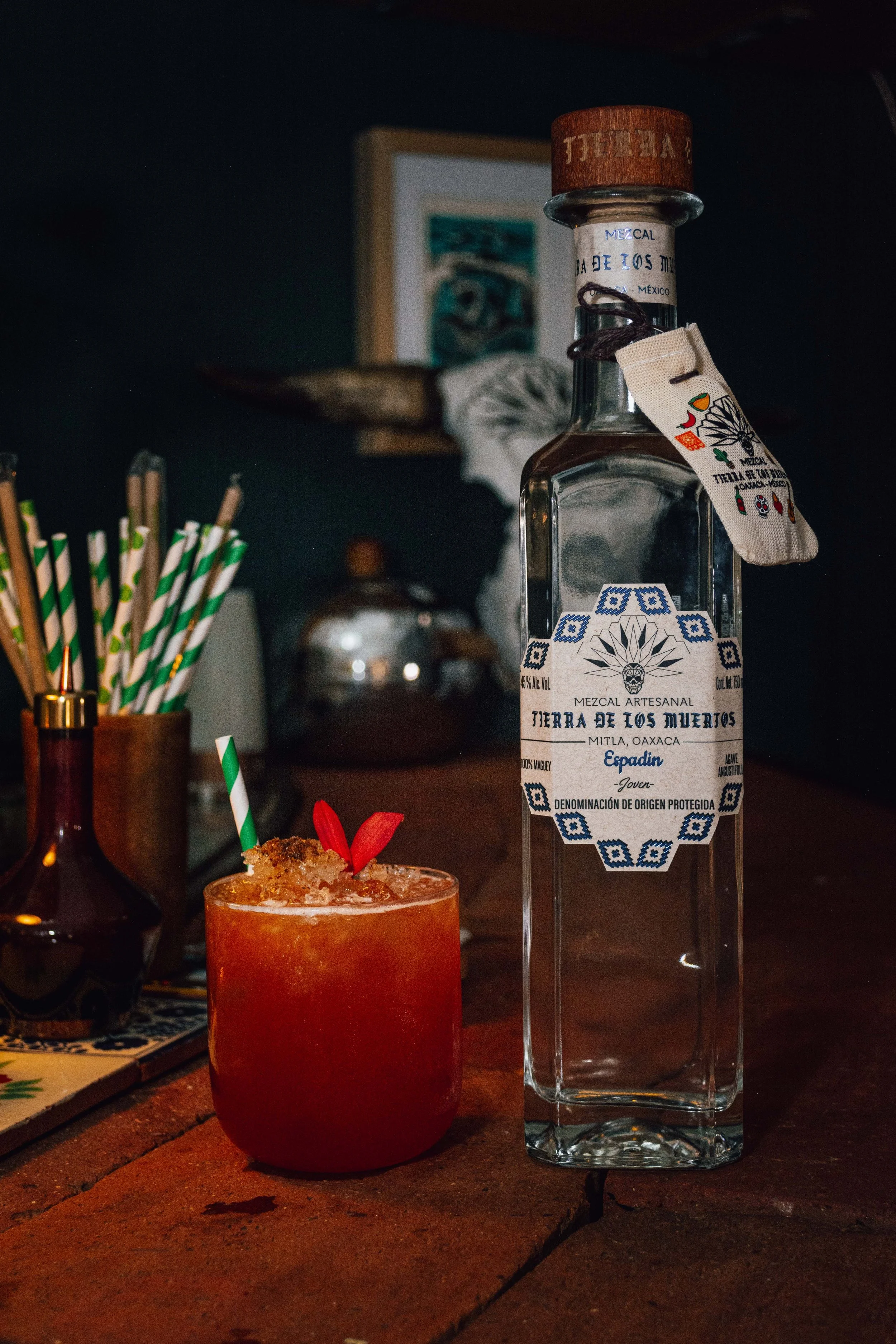 A bottle of Tierra de Los Muertos mezcal and a red cocktail with a straw, flower garnish, and crushed ice on a wooden bar counter.