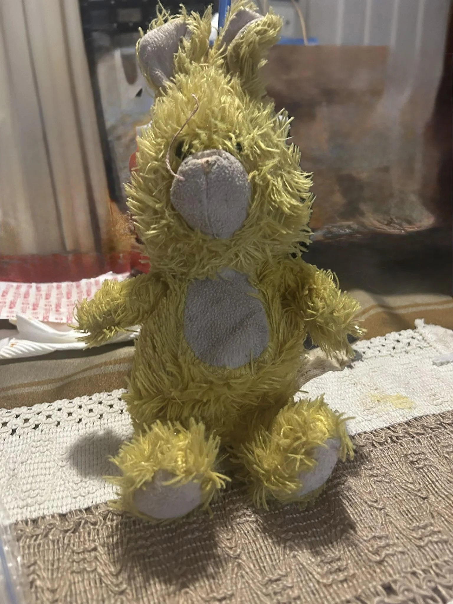 Yellow stuffed animal bear with fuzzy fur and a large nose sitting on a woven cloth surface.