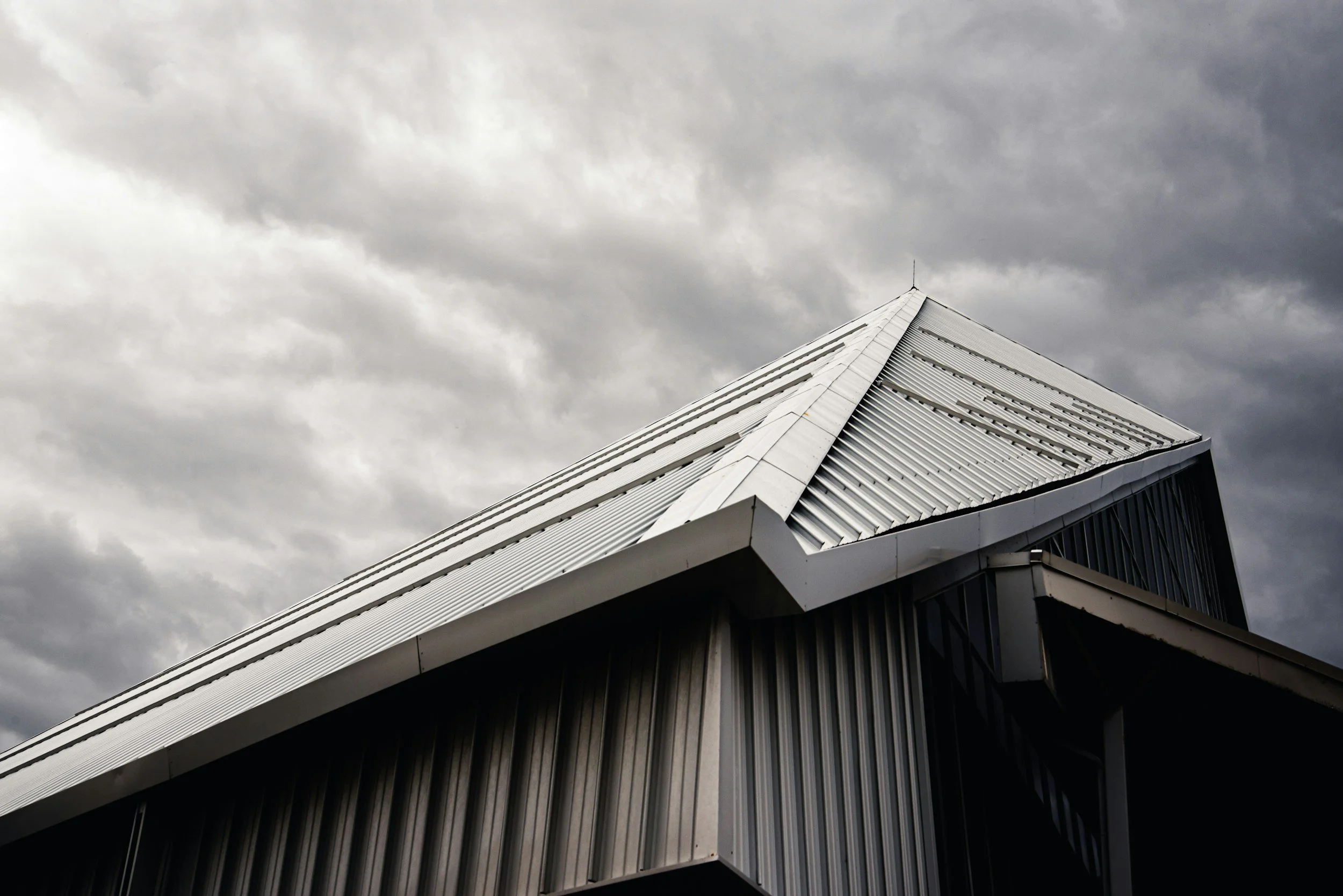 A modern building with metallic siding and a sloped, angular roof against a cloudy sky.