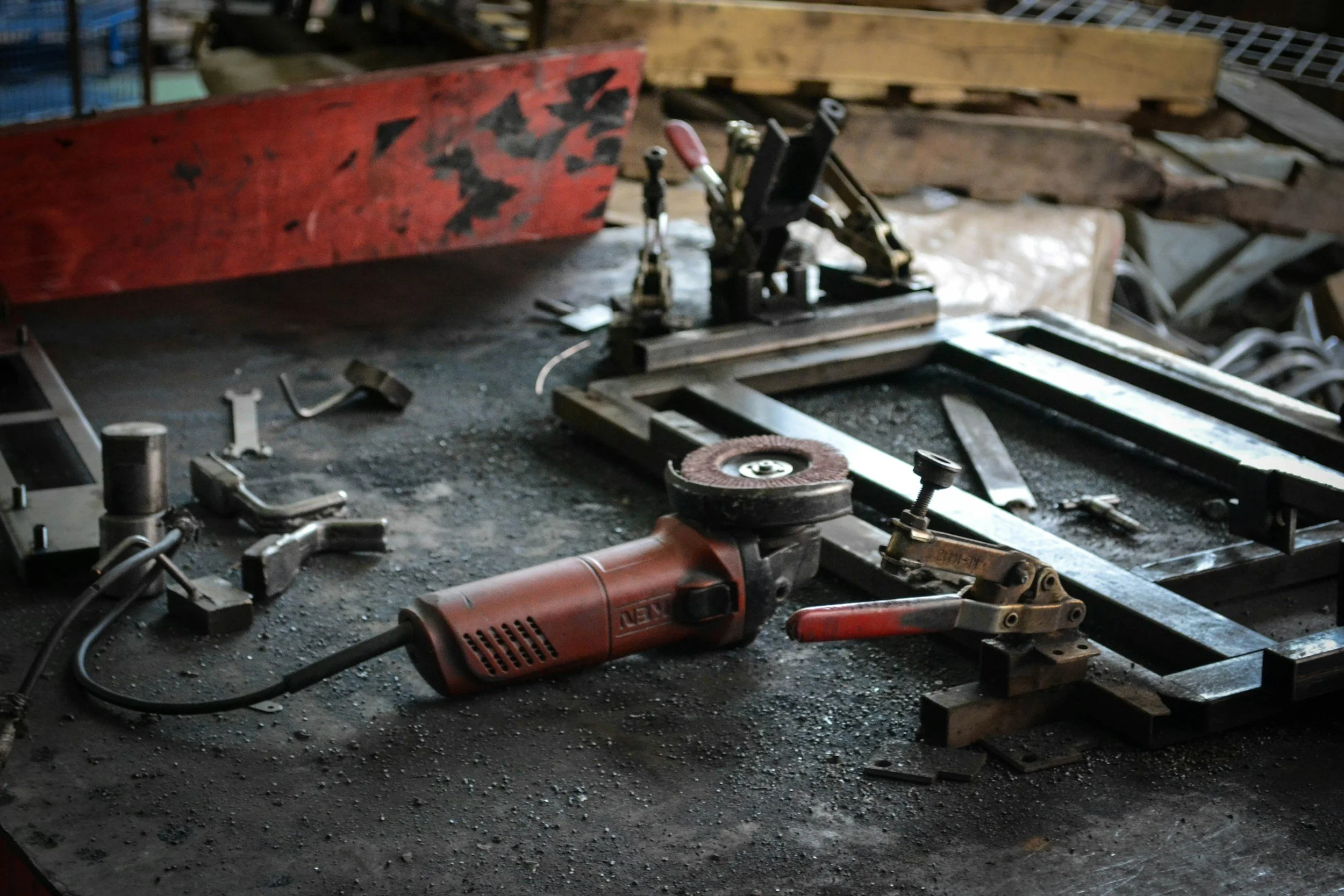 A cluttered workshop workbench with a red angle grinder, a frame, and various tools and materials scattered around.