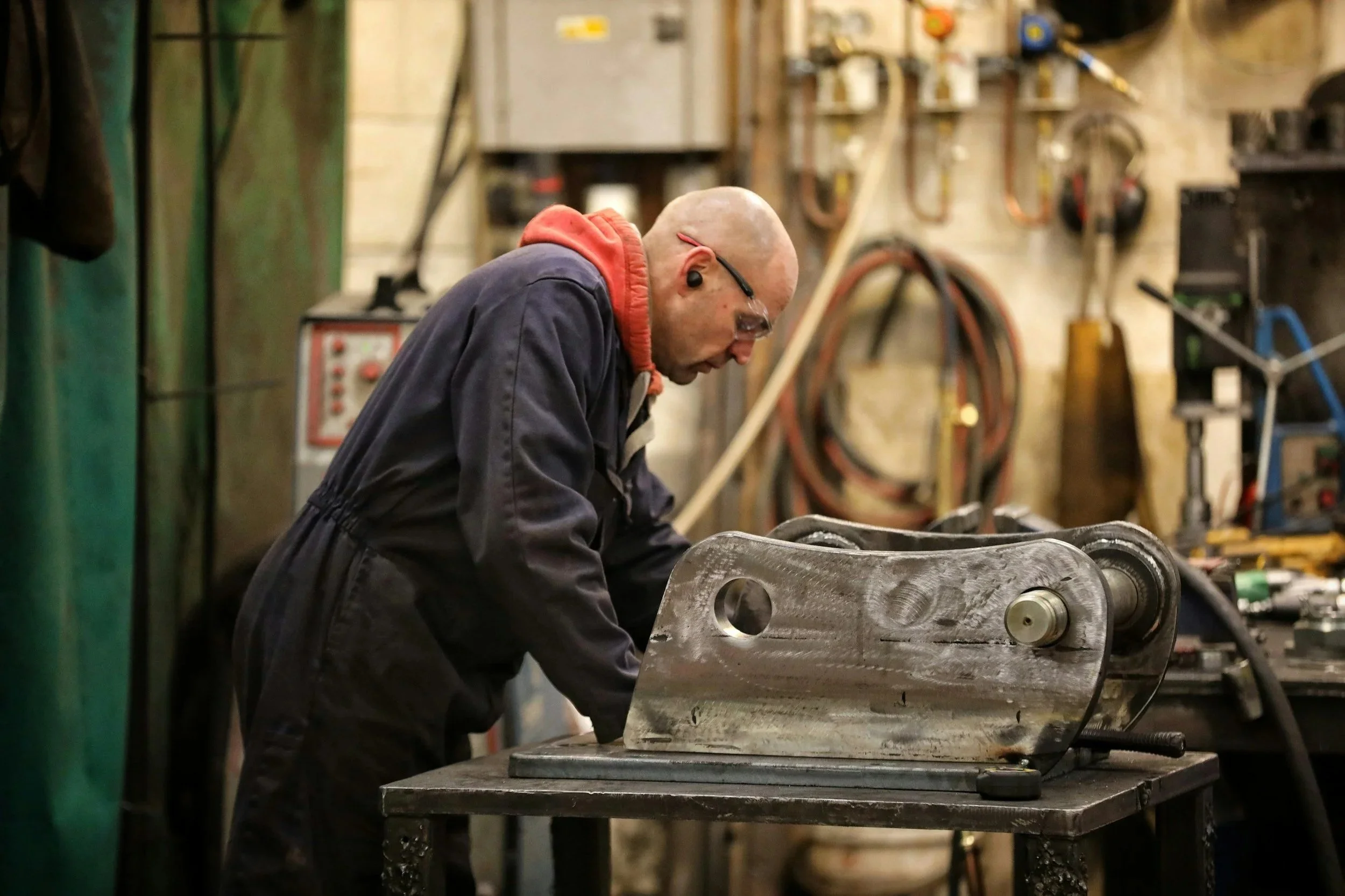 A man in safety glasses and ear protection working on a metal machine part in a workshop.
