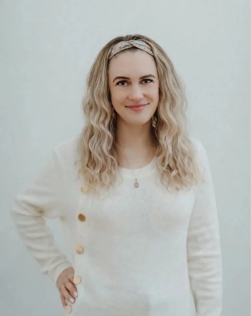 A woman with shoulder-length curly blonde hair, wearing a light-colored headband and a cream-colored top with buttons on the side, smiling at the camera against a plain white background.