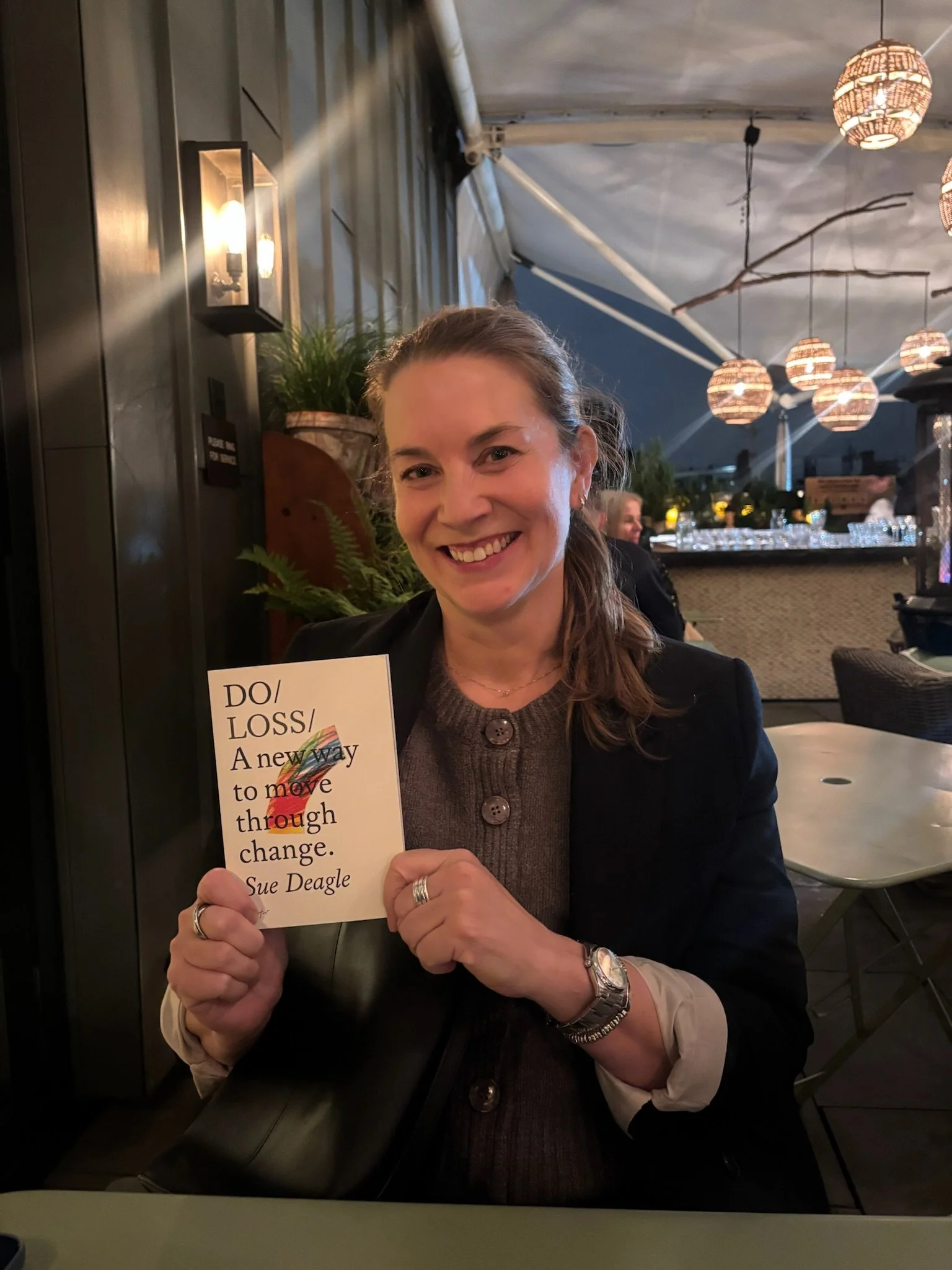A woman (Sue Deagle) smiling and holding her book titled 'DO LOSS: A new way to move through change' by Sue Deagle, in a restaurant with hanging lights and plants in the background.