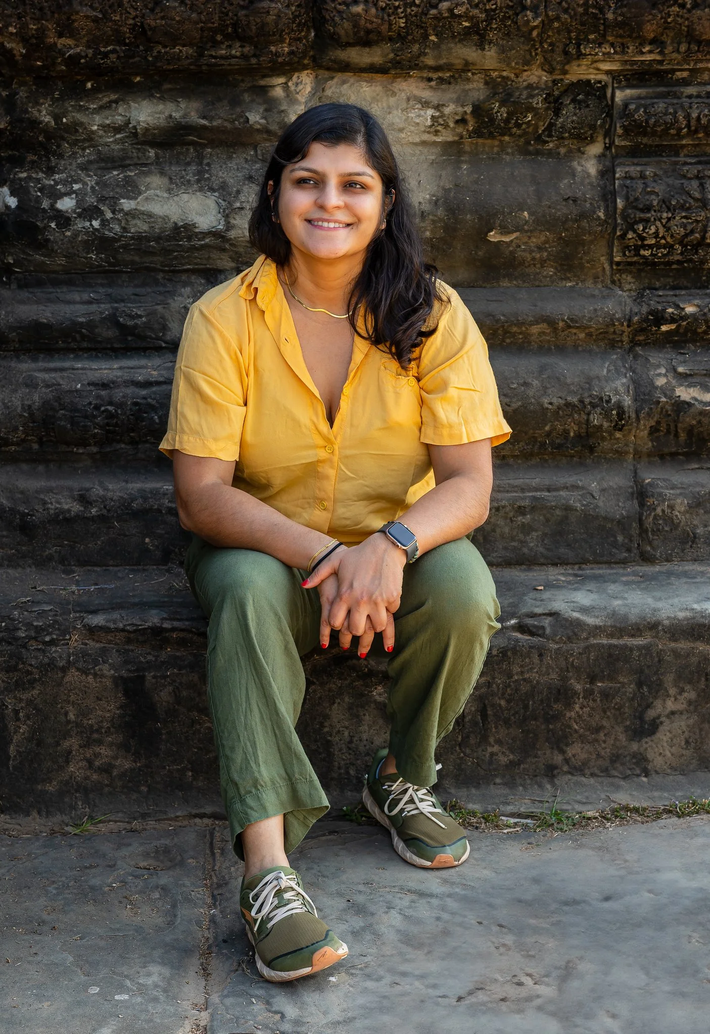 Dr Smriti Singh sitting on stone steps outdoors, wearing a yellow blouse, olive green pants, and sneakers, smiling, with dark hair and a smartwatch on her wrist, in front of a dark stone wall.