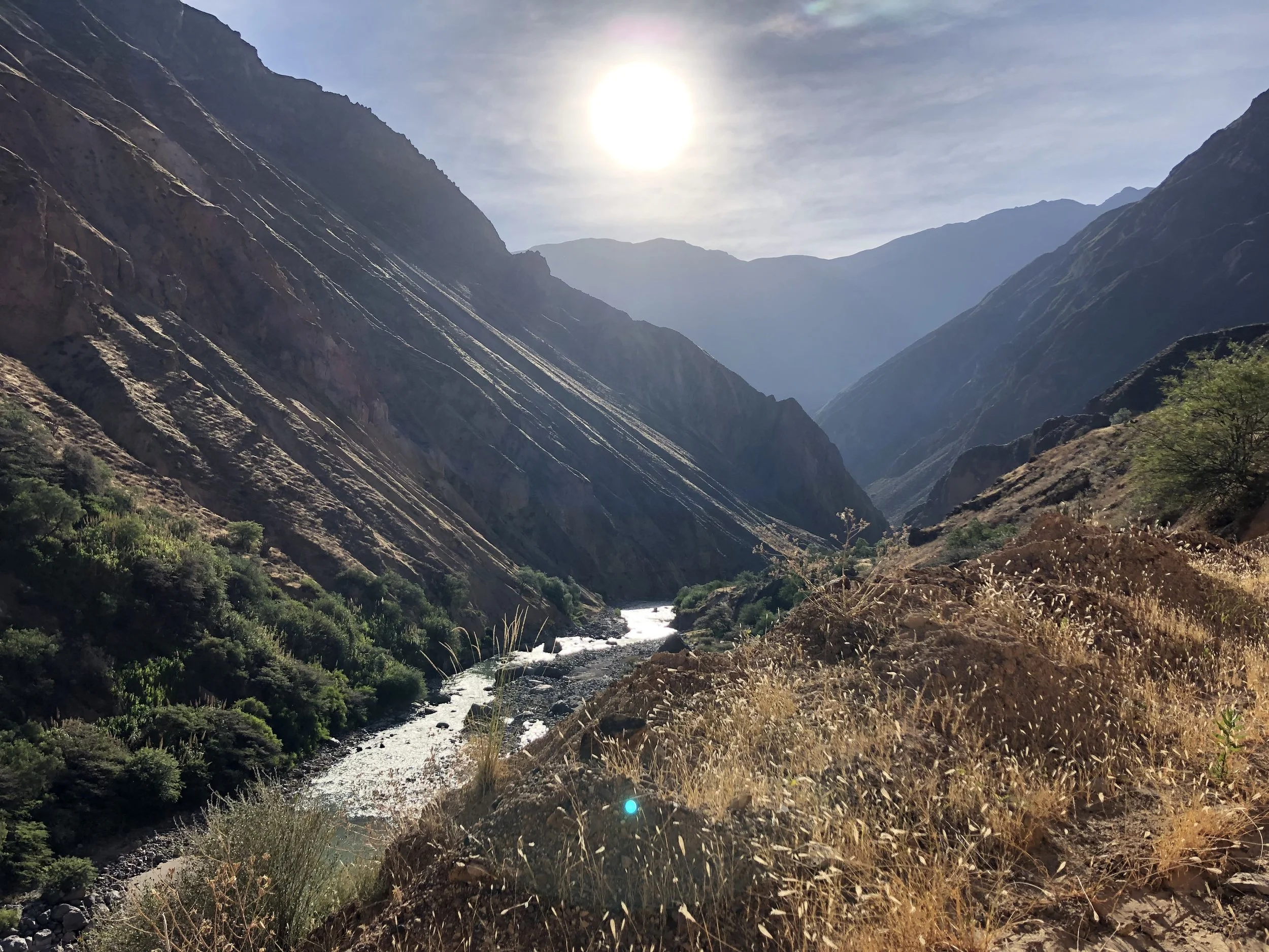 A mountainous landscape with a river flowing through a valley, surrounded by steep slopes with sparse vegetation, under a bright sun in a partly cloudy sky.