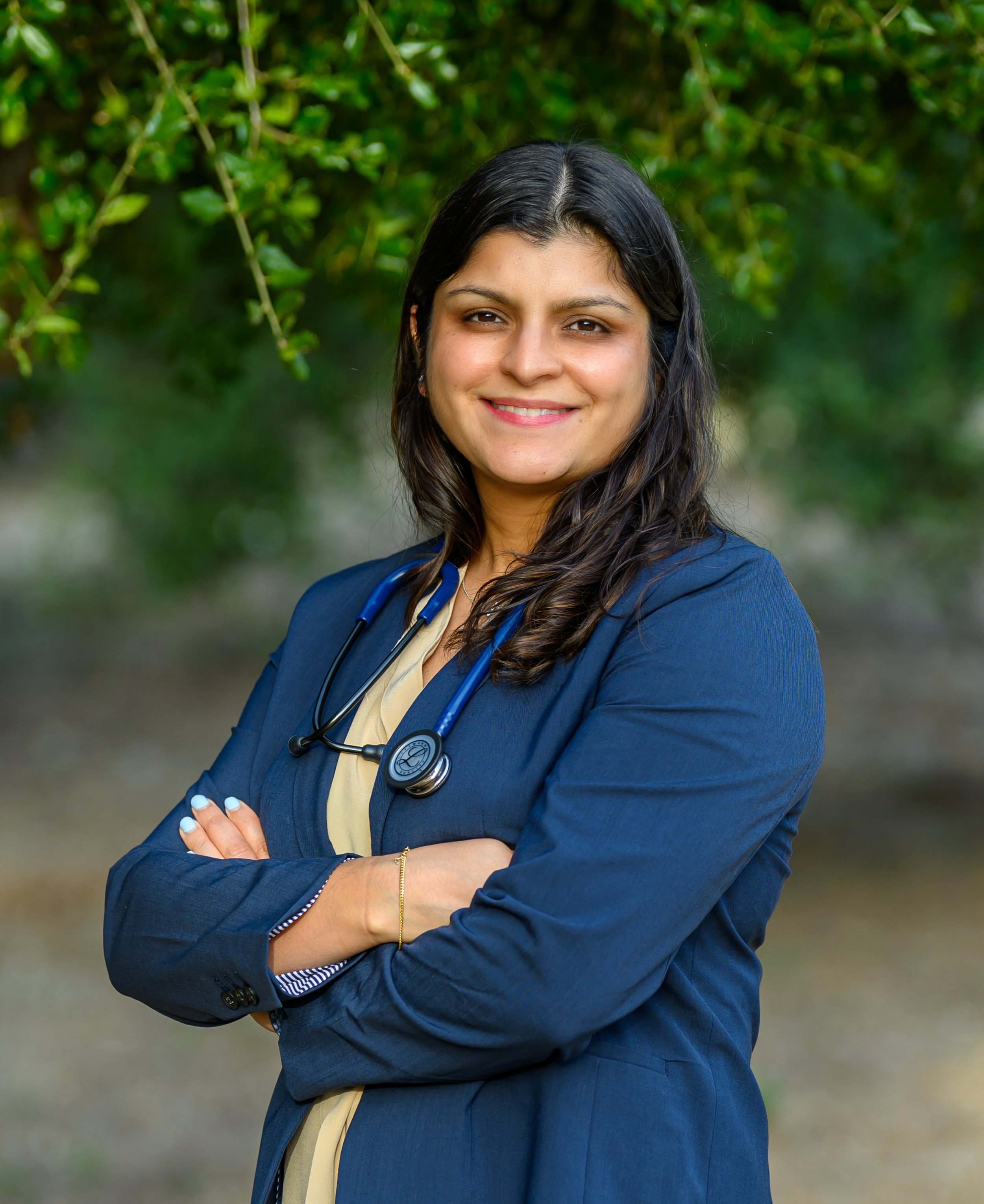 Image of Dr Smriti Singh, a doctor who provides health and wellness services, with dark hair wearing a navy blue blazer, a medical stethoscope around her neck, and a yellow scarf, standing outdoors in front of greenery, smiling with crossed arms.