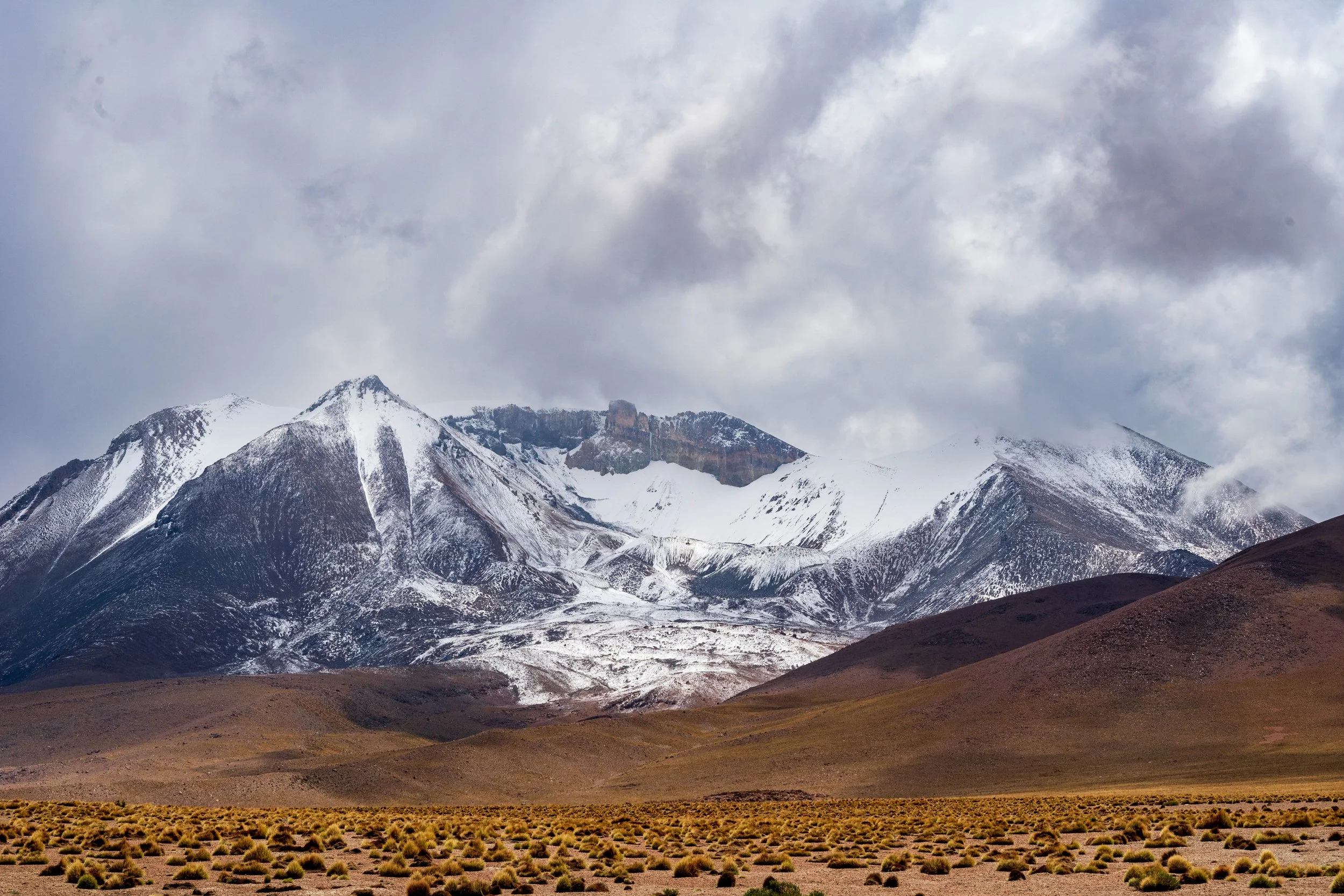 Snow-capped mountain range under cloudy sky with arid plain and sparse vegetation in foreground. Trailhead wellness