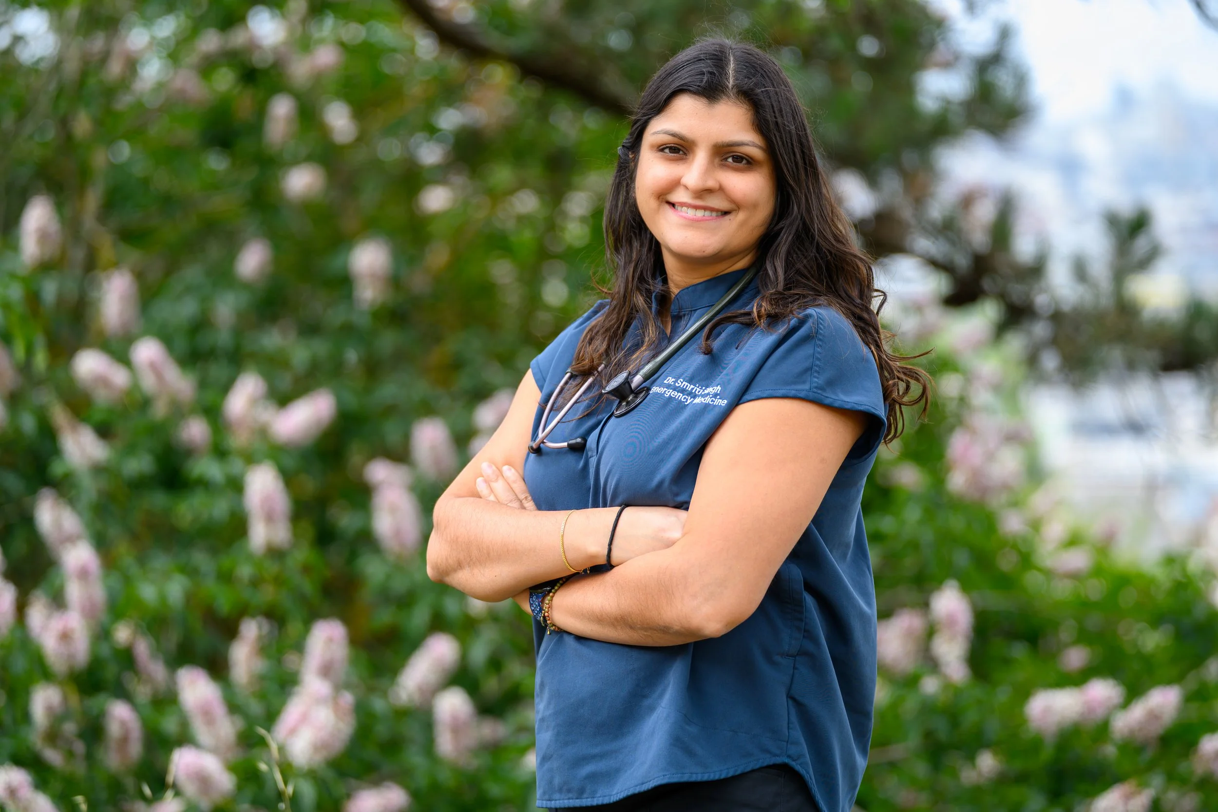Dr Smriti Singh, doctor at Trailhead Wellness, wearing medical scrubs stands outdoors with arms crossed, smiling, in front of a blurred background of trees and pink flowers.