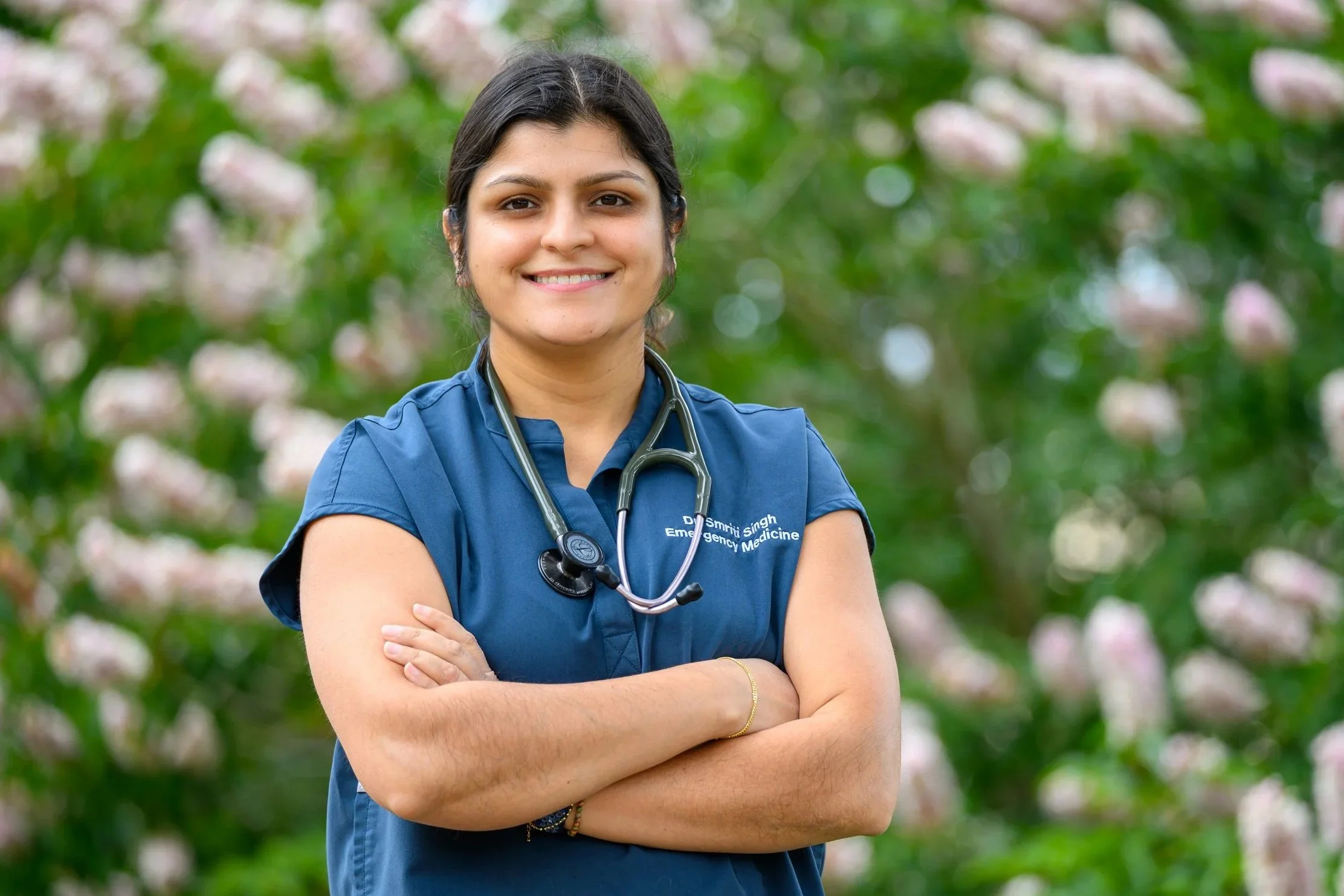 A female healthcare professional with dark hair wearing blue scrubs and a stethoscope around her neck, standing outdoors with her arms crossed and smiling, with pink and white flowers in the background.