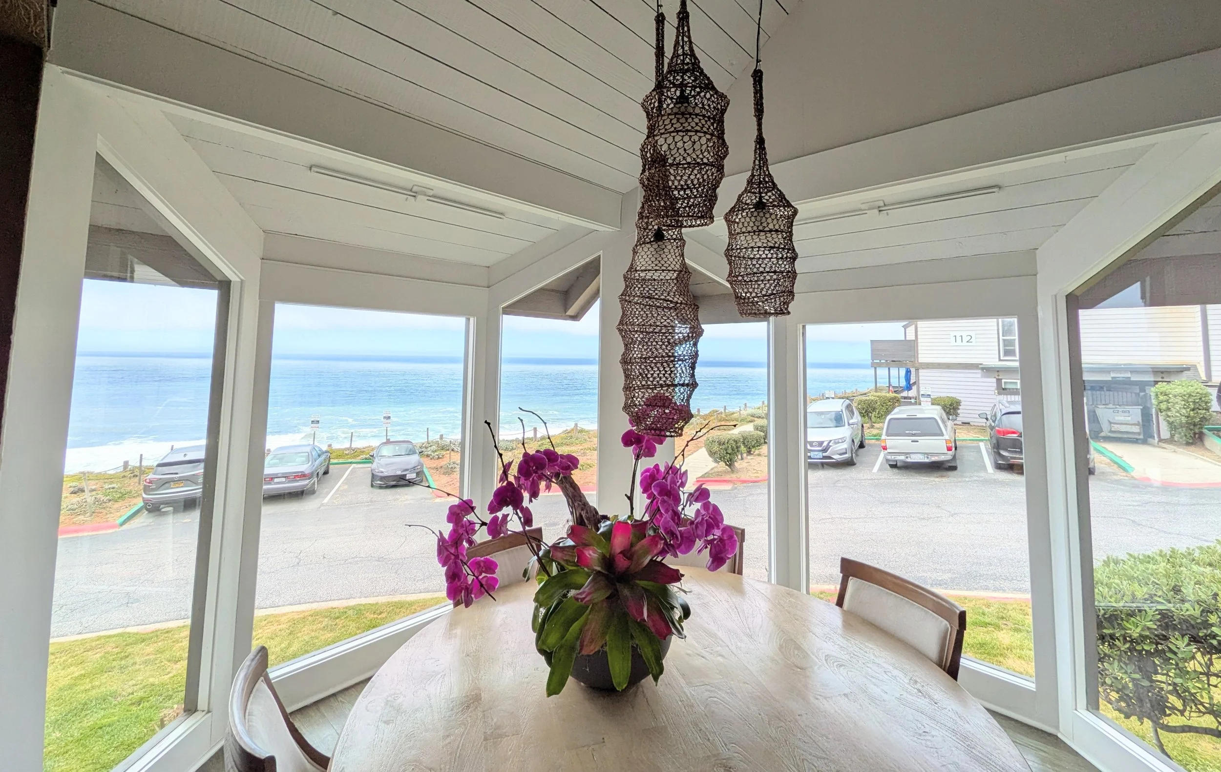 Dining area with a round wooden table decorated with pink orchids, large just cleaned windows showing parking lot and ocean view, woven pendant lights hanging from ceiling.