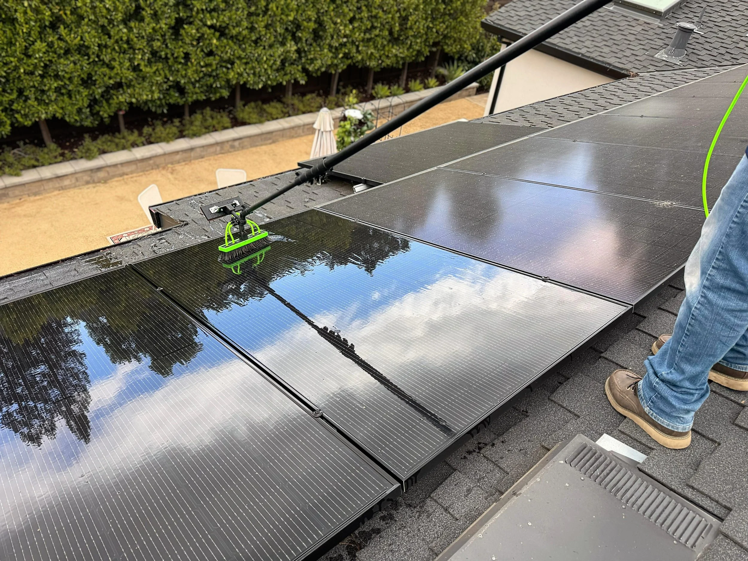 Worker cleaning the surface of solar panels on a sloped roof with a brush and hose.