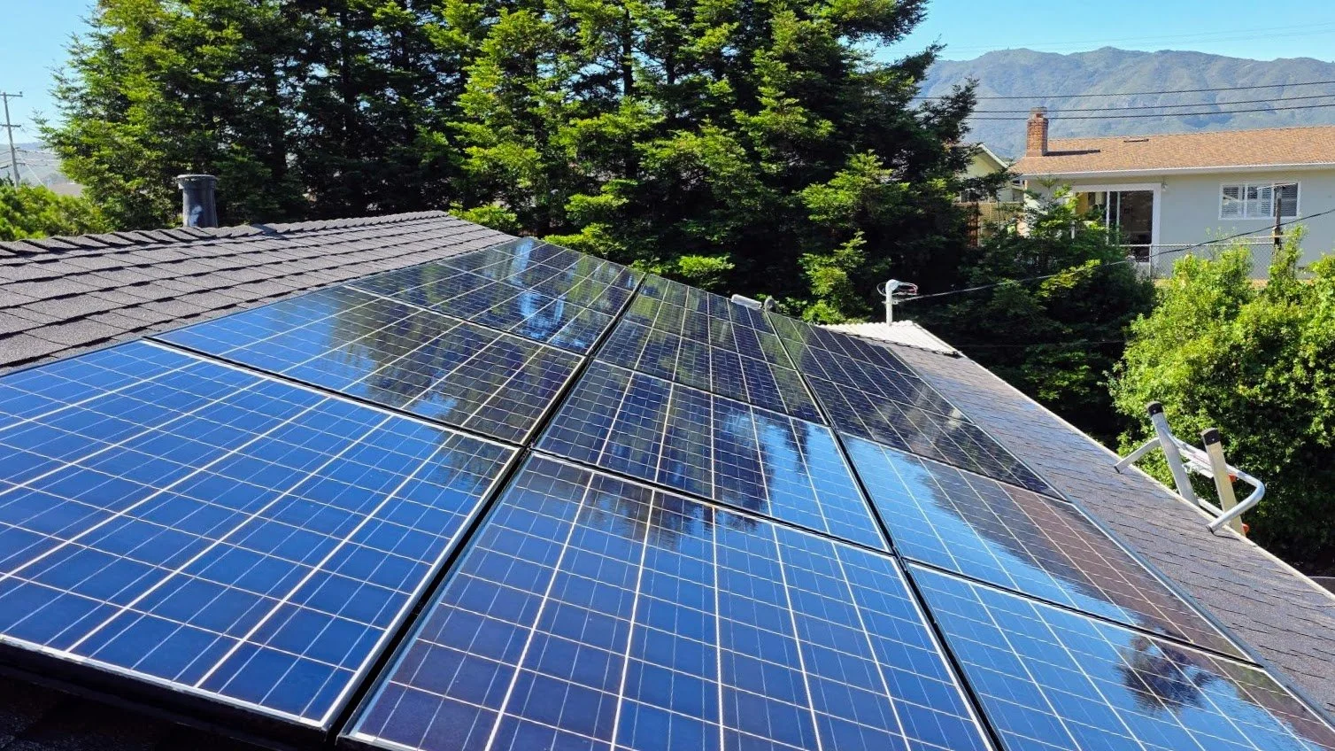 Solar panels installed on a house roof, surrounded by trees, with houses and mountains in the background.