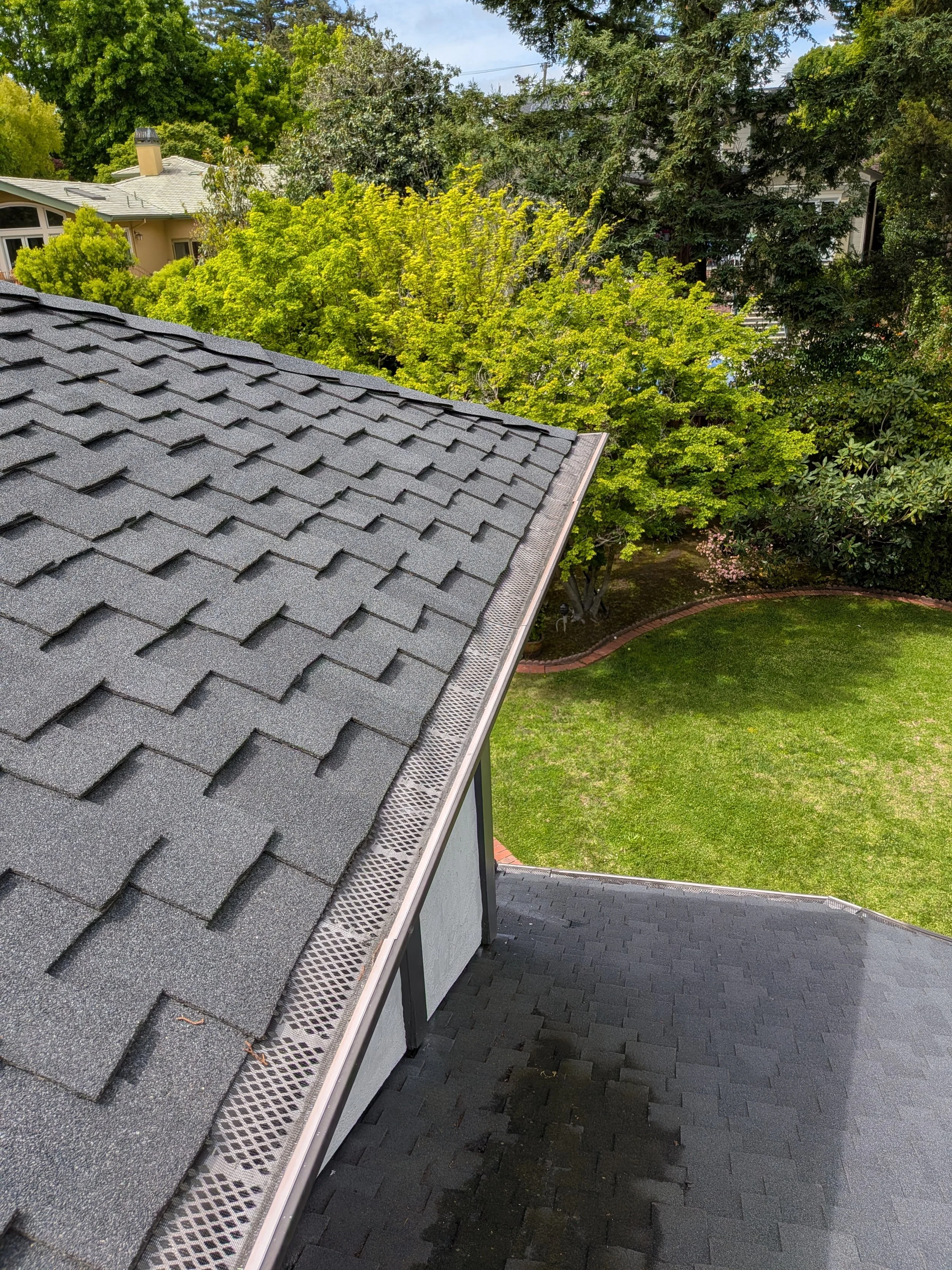 A rooftop with dark gray shingles, surrounded by trees with green foliage, a lawn, and a brick border in a residential area.