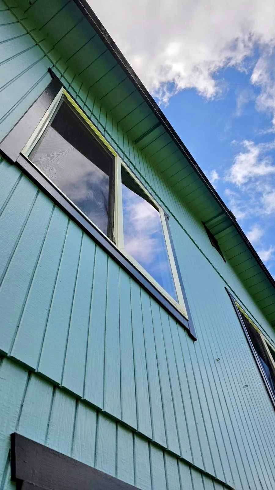 Low angle view of a turquoise-colored house exterior with large pristine windows, under a partly cloudy sky.