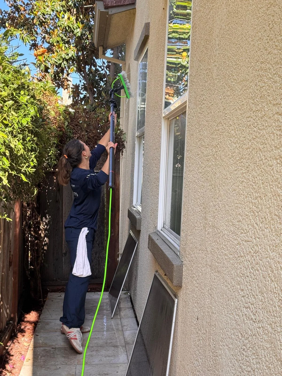 A woman cleaning windows on the exterior of a house with a long-handled scrub brush.