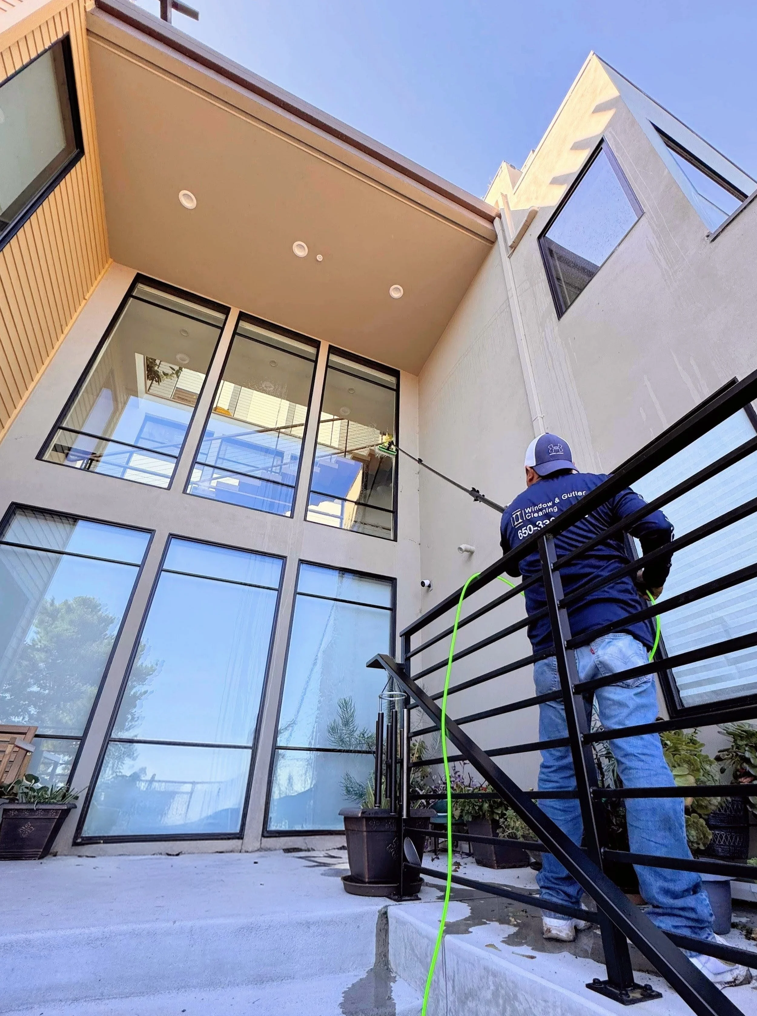 A worker in a blue shirt and jeans is pressure washing the outdoor concrete patio of a modern multi-story house with large glass windows. The house has a beige and brown exterior with a combination of wood and stucco finishes. The worker is standing 
