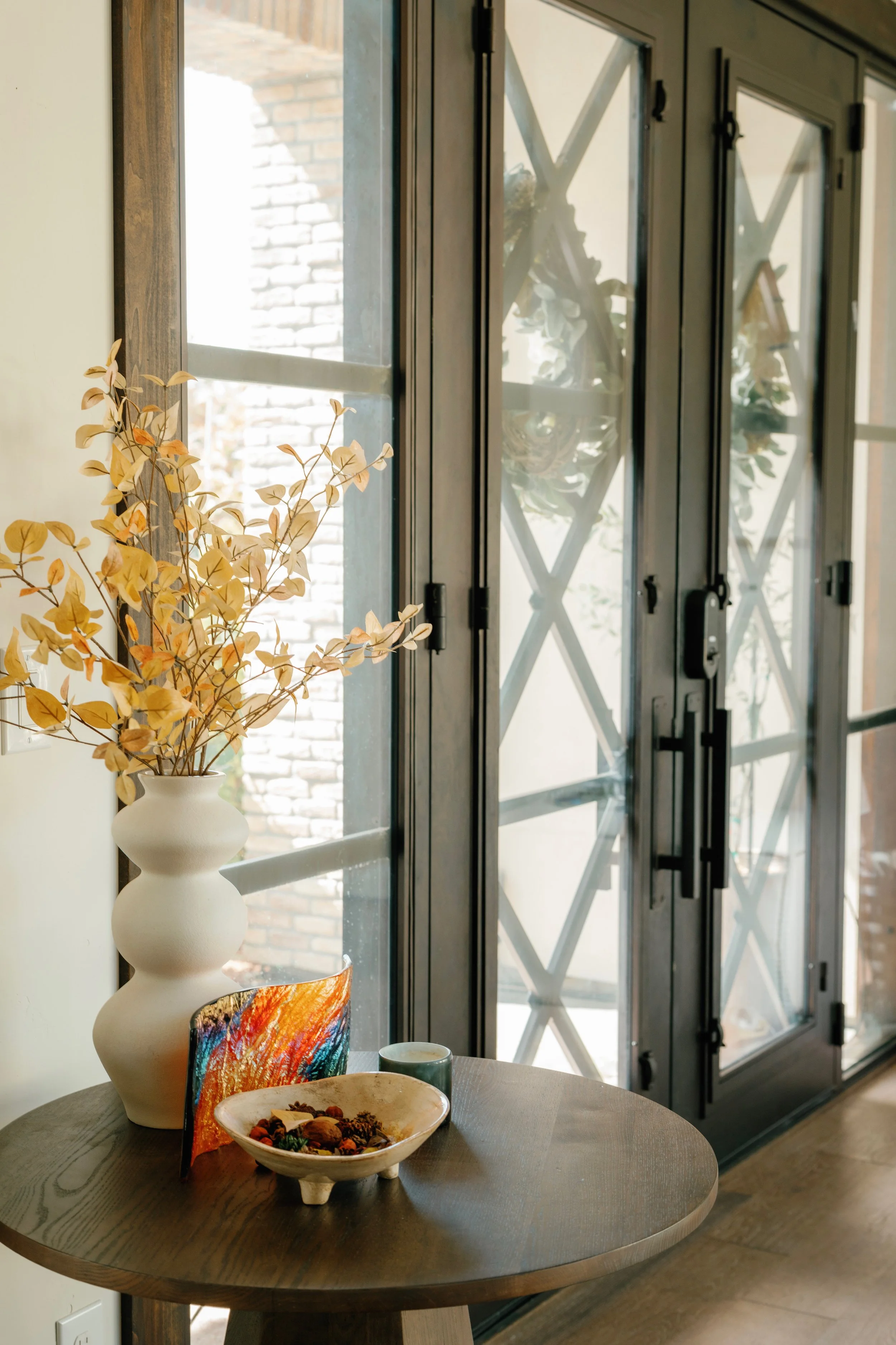 Round wooden table with a decorative bowl containing dried flowers or potpourri, a small teal-colored cup or container, and a colorful abstract art piece, beside a white vase with dried branches, near a large window with black metal framing and crisscross grid pattern, letting in natural light.