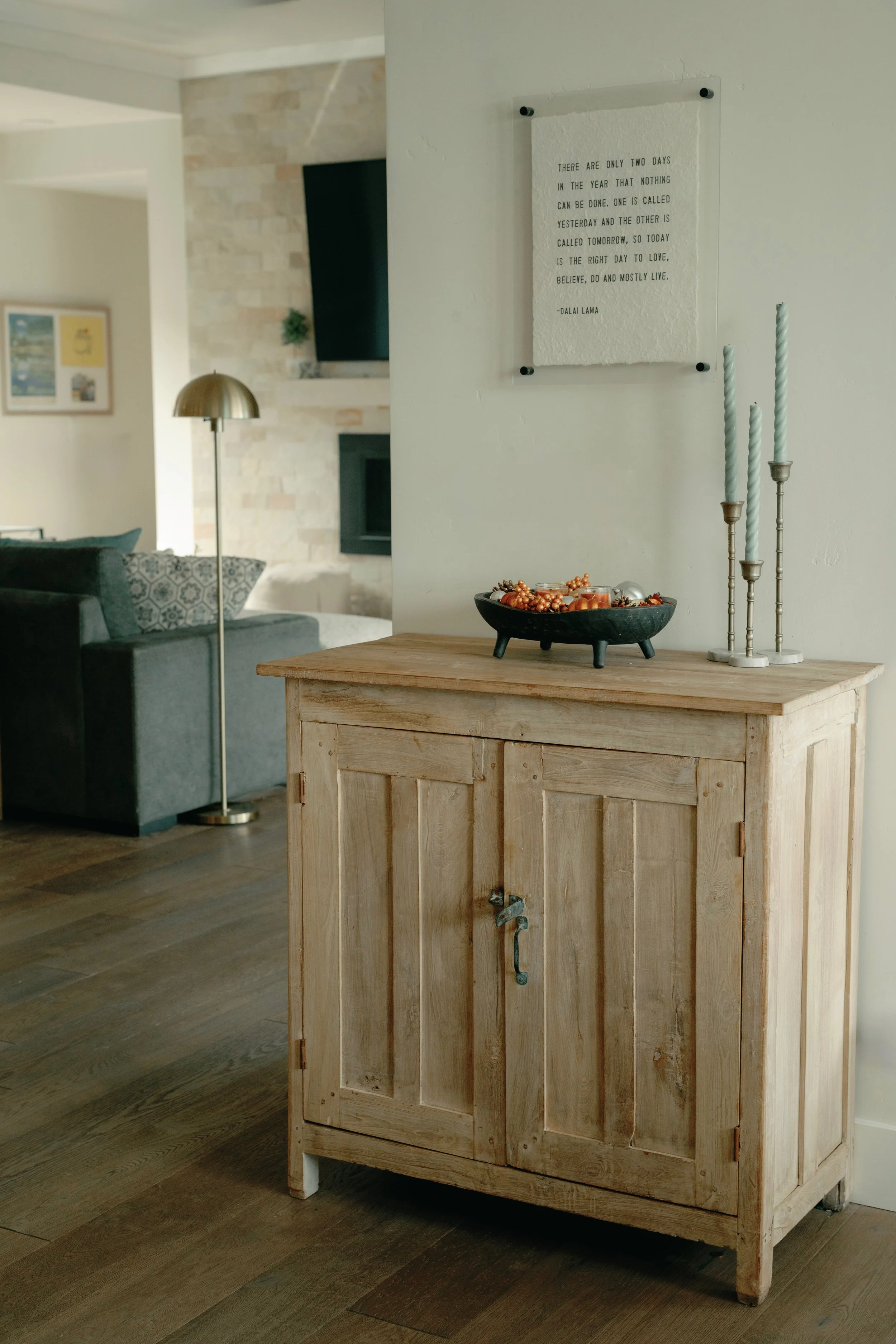 A wooden cabinet with candle holders and a decorative bowl on top, against a white wall with a quote hanging above, in a living room with a sofa and a floor lamp.