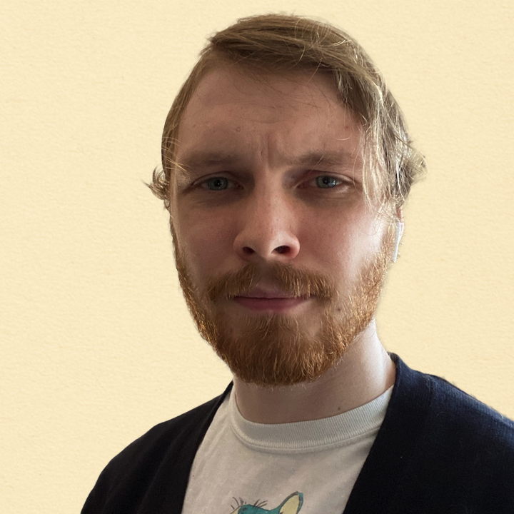 A young man with red hair and a beard looking directly at the camera against a plain beige background.