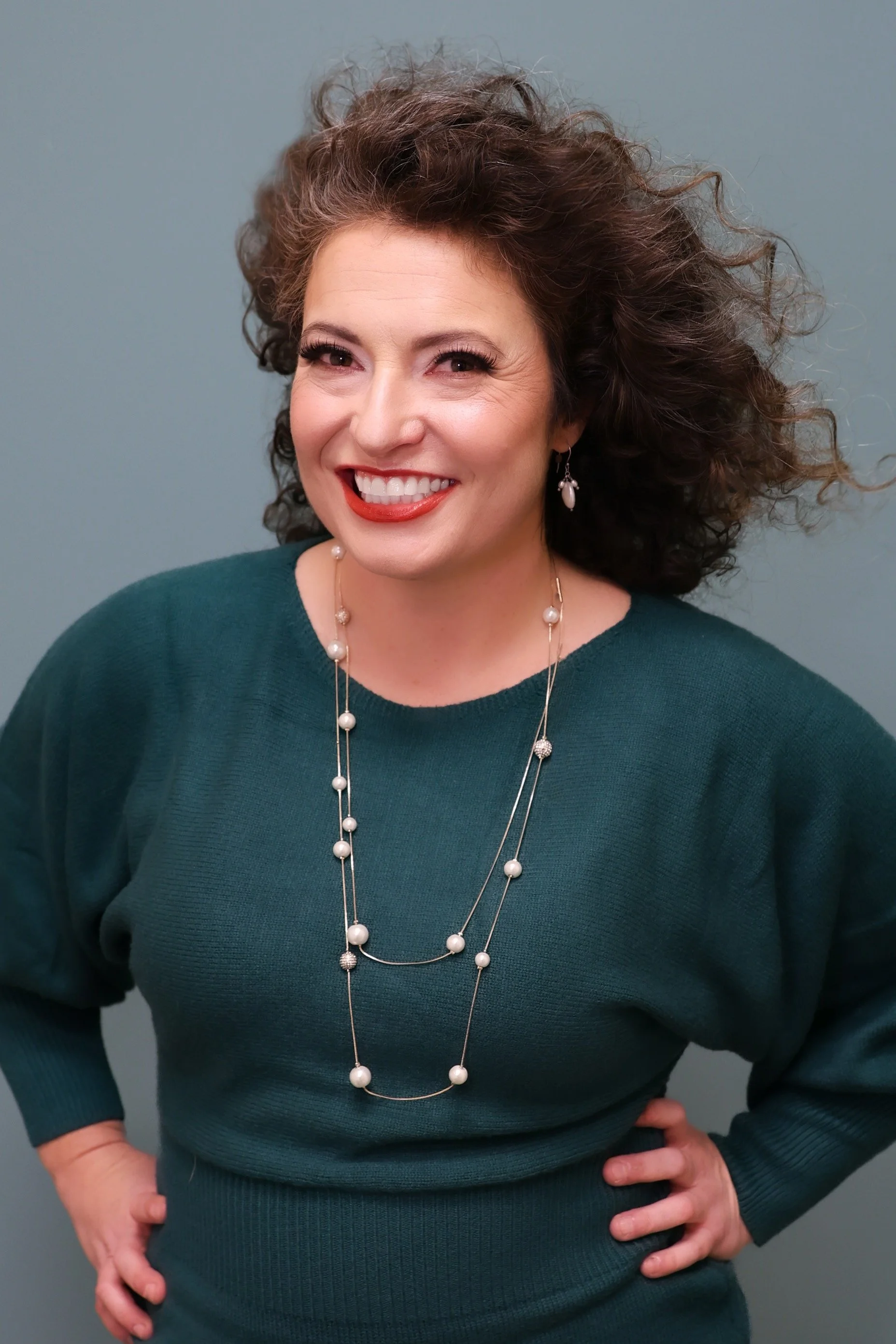 Smiling woman with curly hair wearing a teal sweater and pearl necklace, standing with hands on hips against a neutral gray background.
