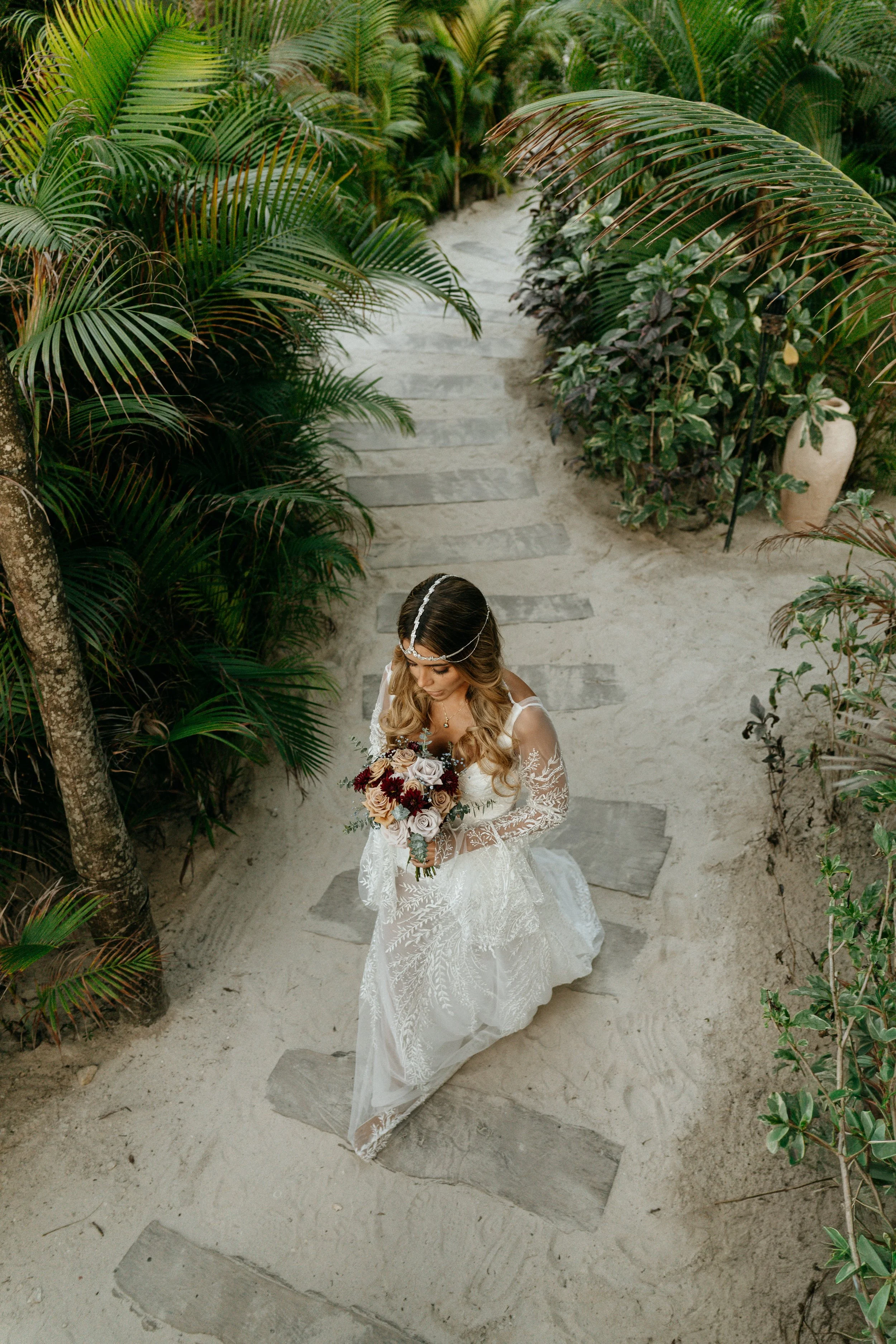 Bride walking through a tropical setting in Holbox while holding a bouquet of roses and greenery.