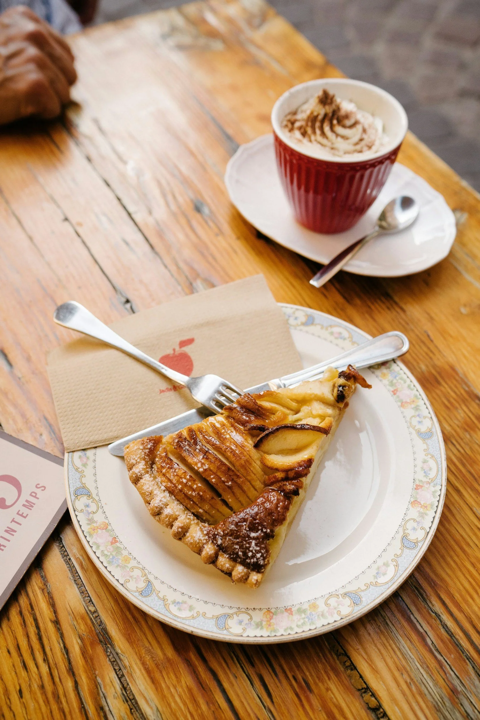 A slice of apple pie on a decorative plate with a fork, and a cup of coffee with whipped cream and cinnamon on a saucer and spoon, on a wooden table.