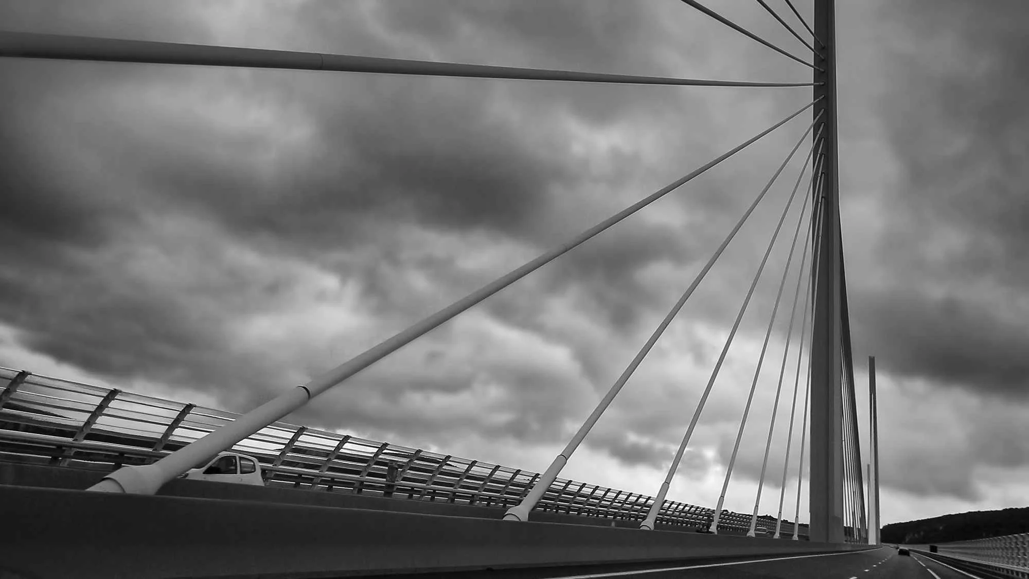 Schwarz-Weiß-Foto eines Brückenpylons mit Kabeln über einer Straße, bei dunklen Wolken am Himmel. In der Auvergne, Frankreich.